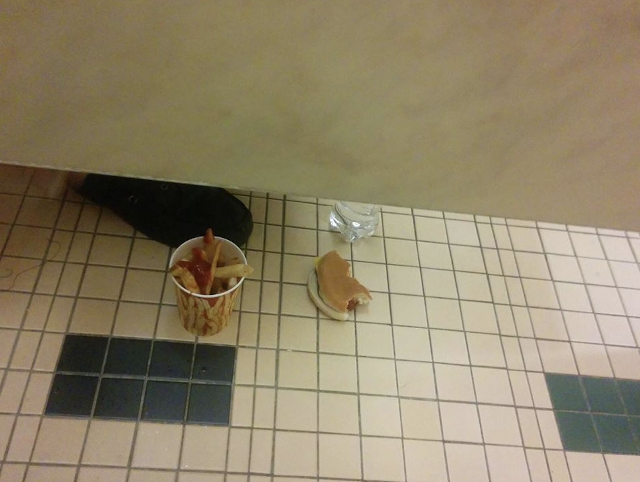 A paper cup of french fries with ketchup and a half-eaten hot dog sit on a tiled restroom floor near a person’s foot.