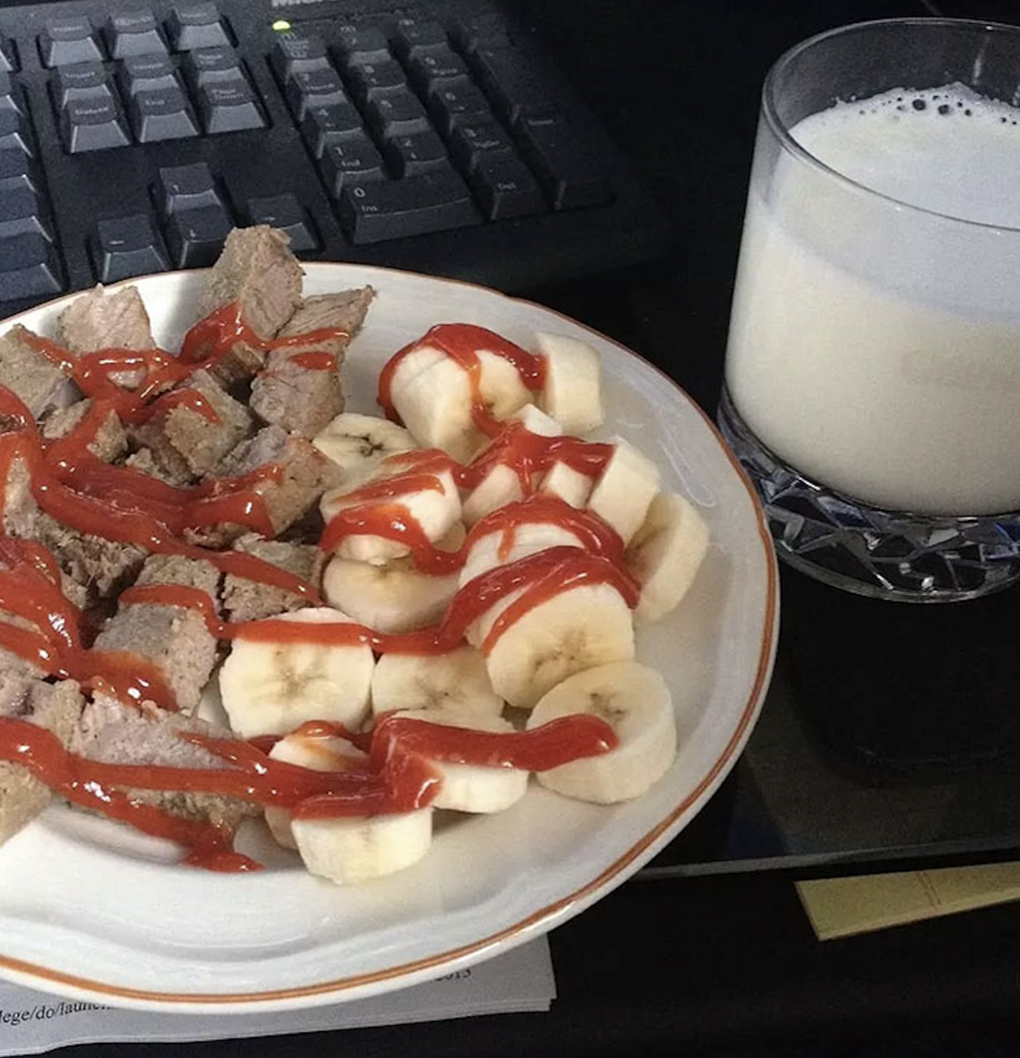 A plate of sliced meat and banana coins drizzled with ketchup, next to a glass of milk and a computer keyboard.