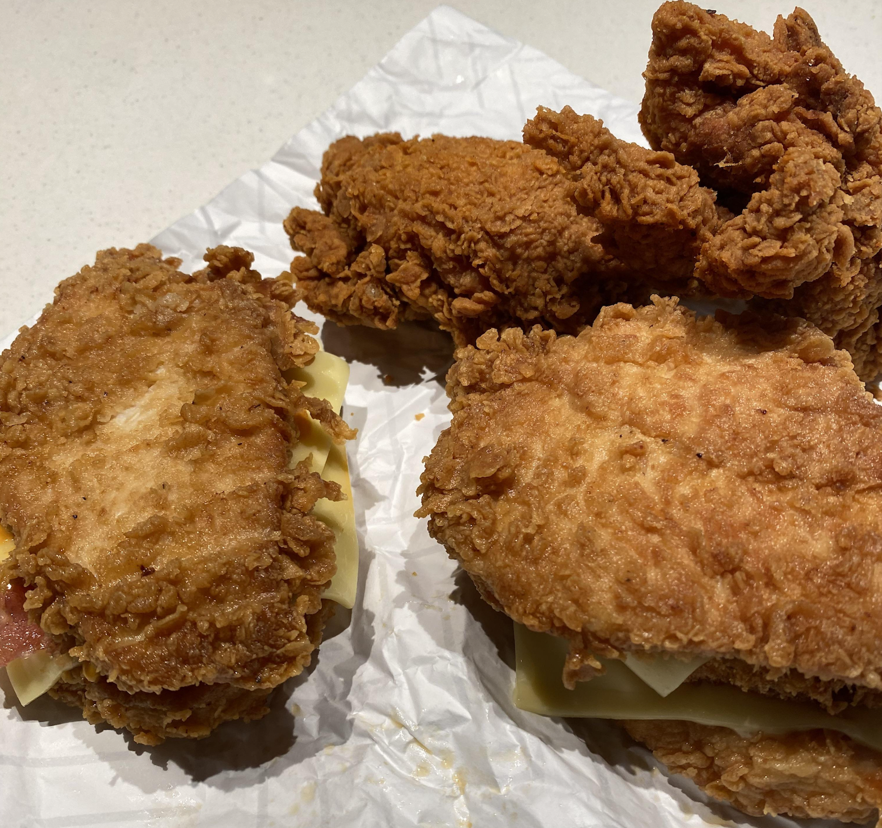 Two fried chicken sandwiches with cheese and a side of fried chicken pieces on parchment paper.