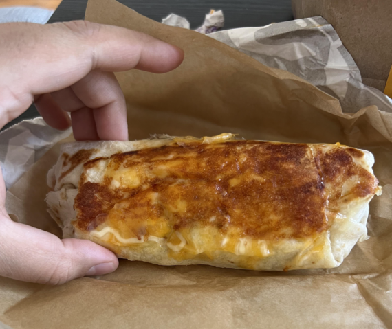 A hand holding a toasted, cheese-crusted burrito wrapped in parchment paper.
