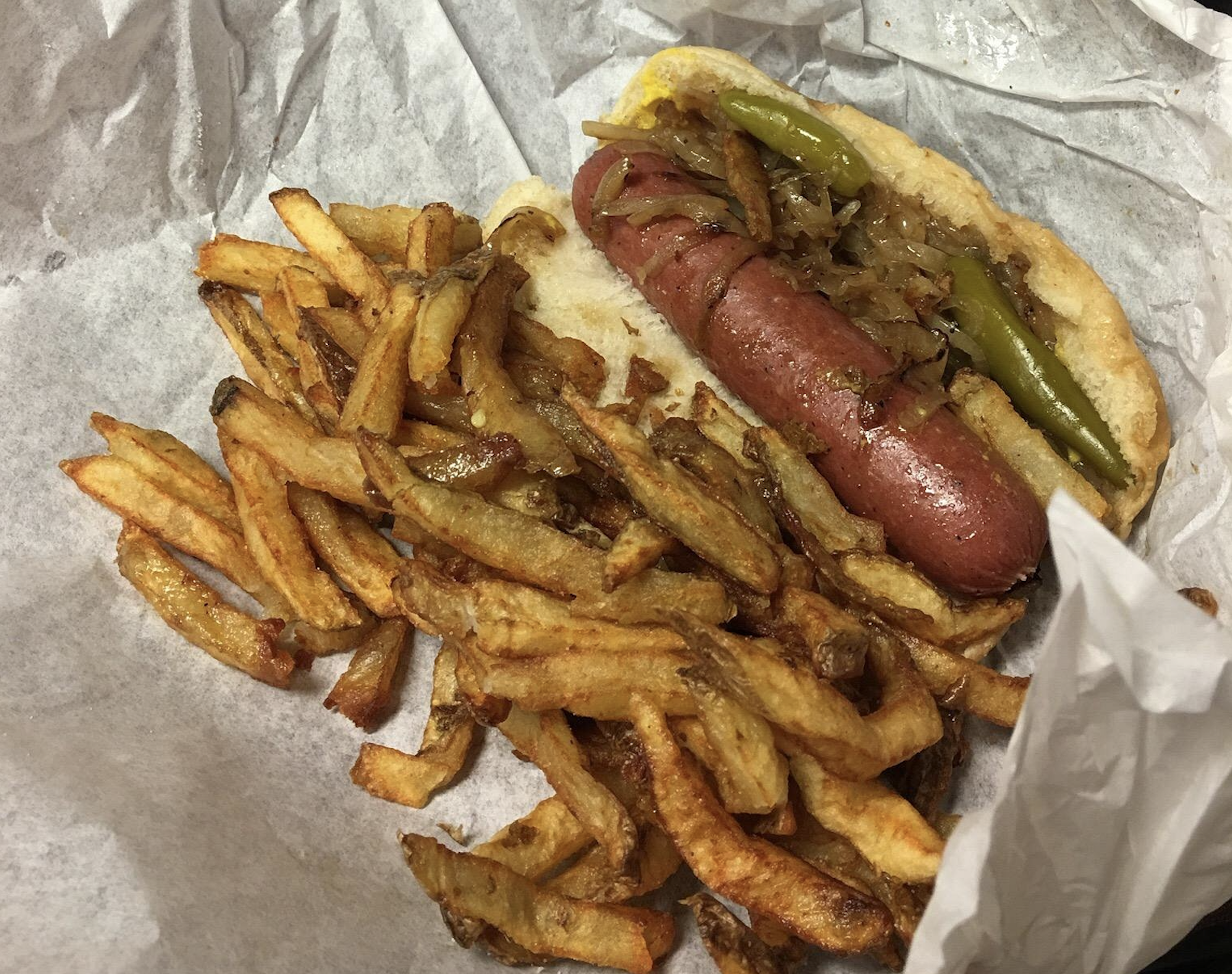 A hot dog topped with sautéed onions and a green chili pepper, served with a pile of thin-cut french fries in a paper-lined basket.