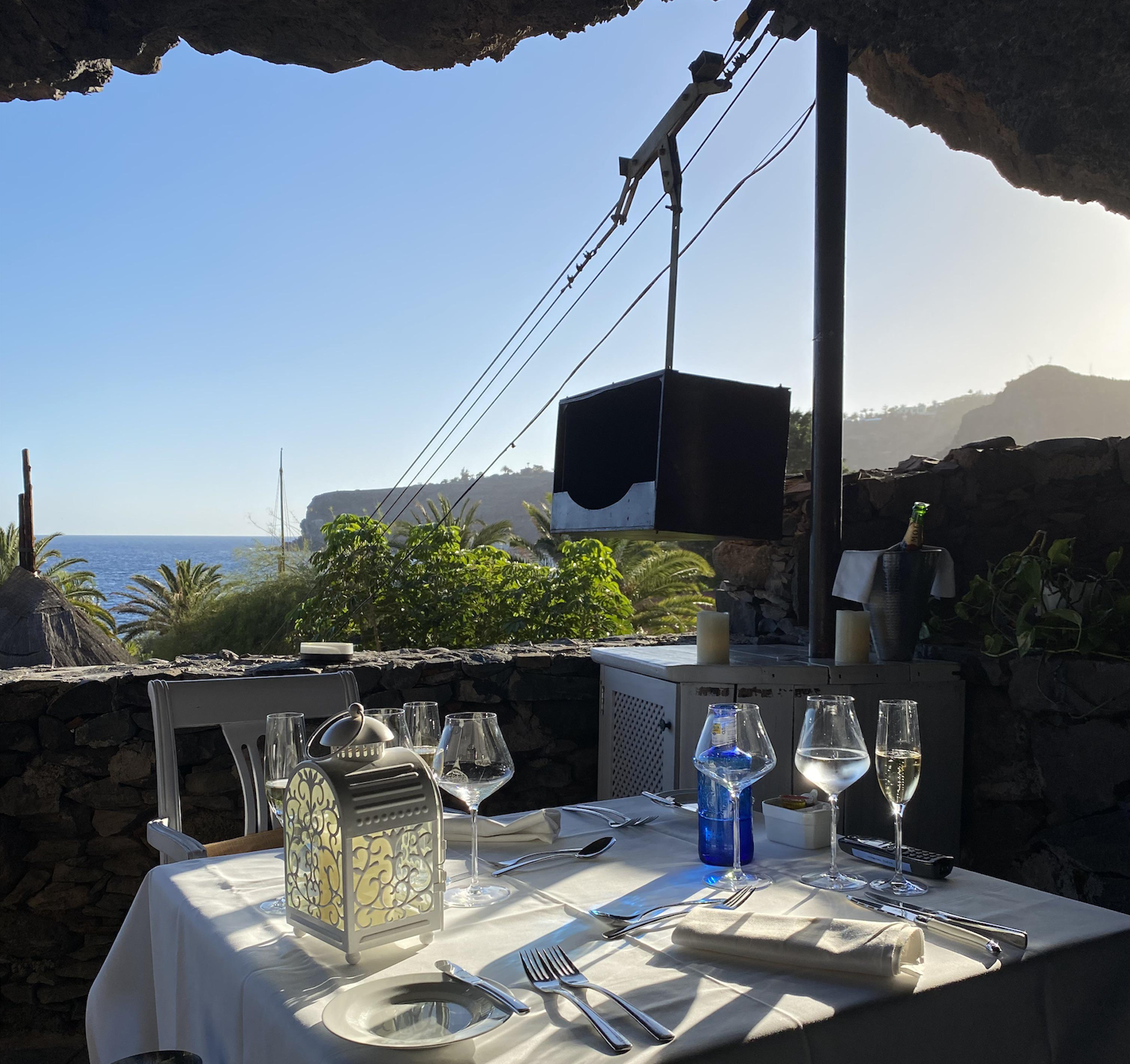A set table with wine glasses, a lantern, and cutlery on a terrace overlooking the ocean under a rock overhang.