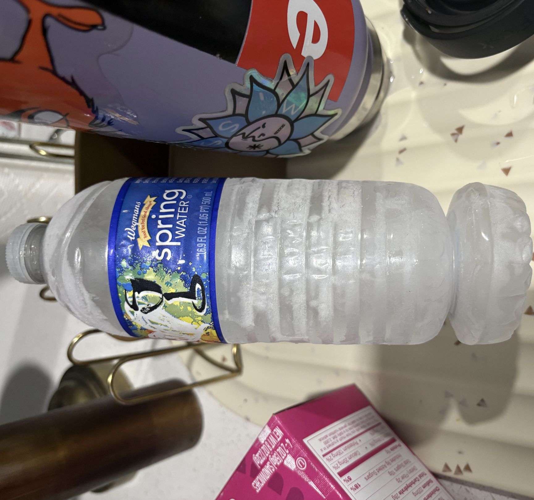 A frosty bottle of Wegmans spring water lying horizontally next to a sticker-covered bottle and a pink box of frozen sandwiches.