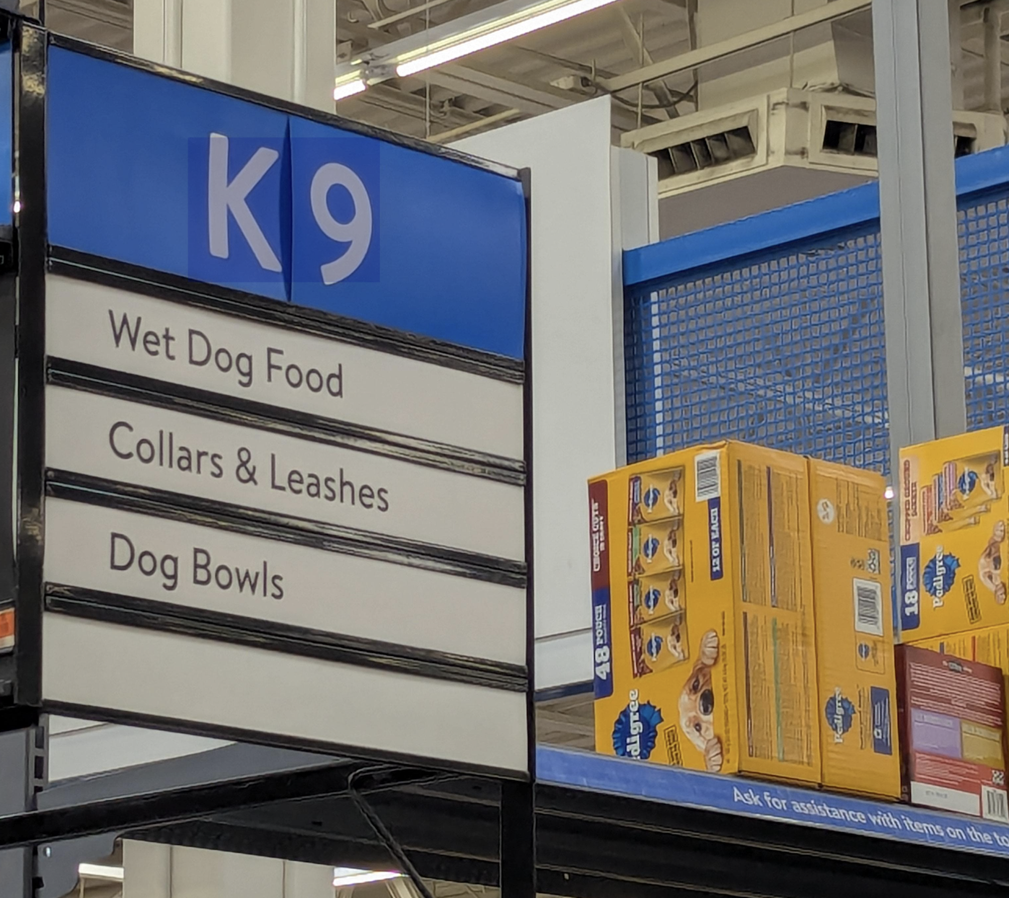 Store aisle sign labeled K9 for wet dog food, collars and leashes, and dog bowls, positioned above shelves stocked with boxes of Pedigree dog food.