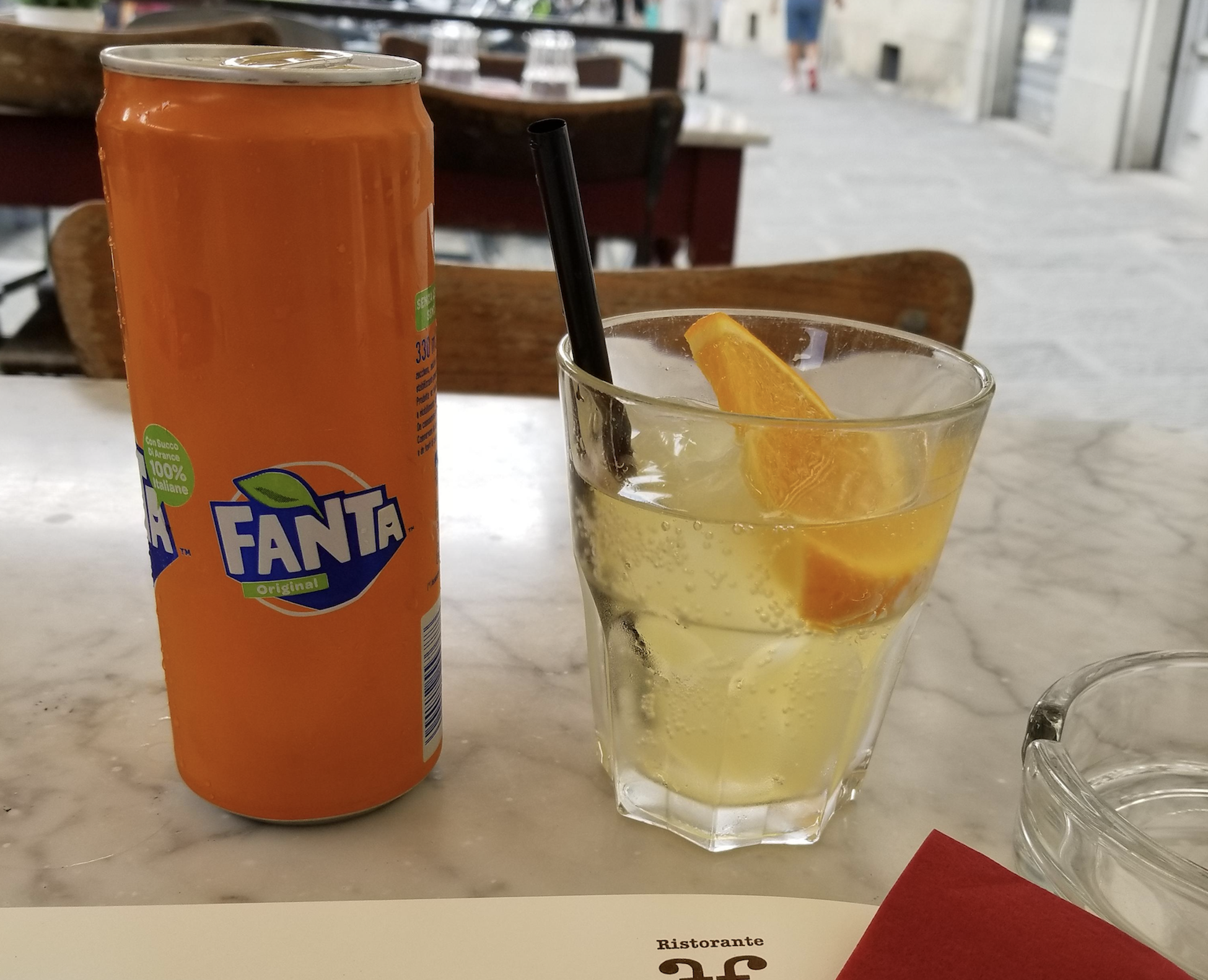 An orange can of Fanta next to a glass of sparkling drink with ice and an orange slice on a café table.