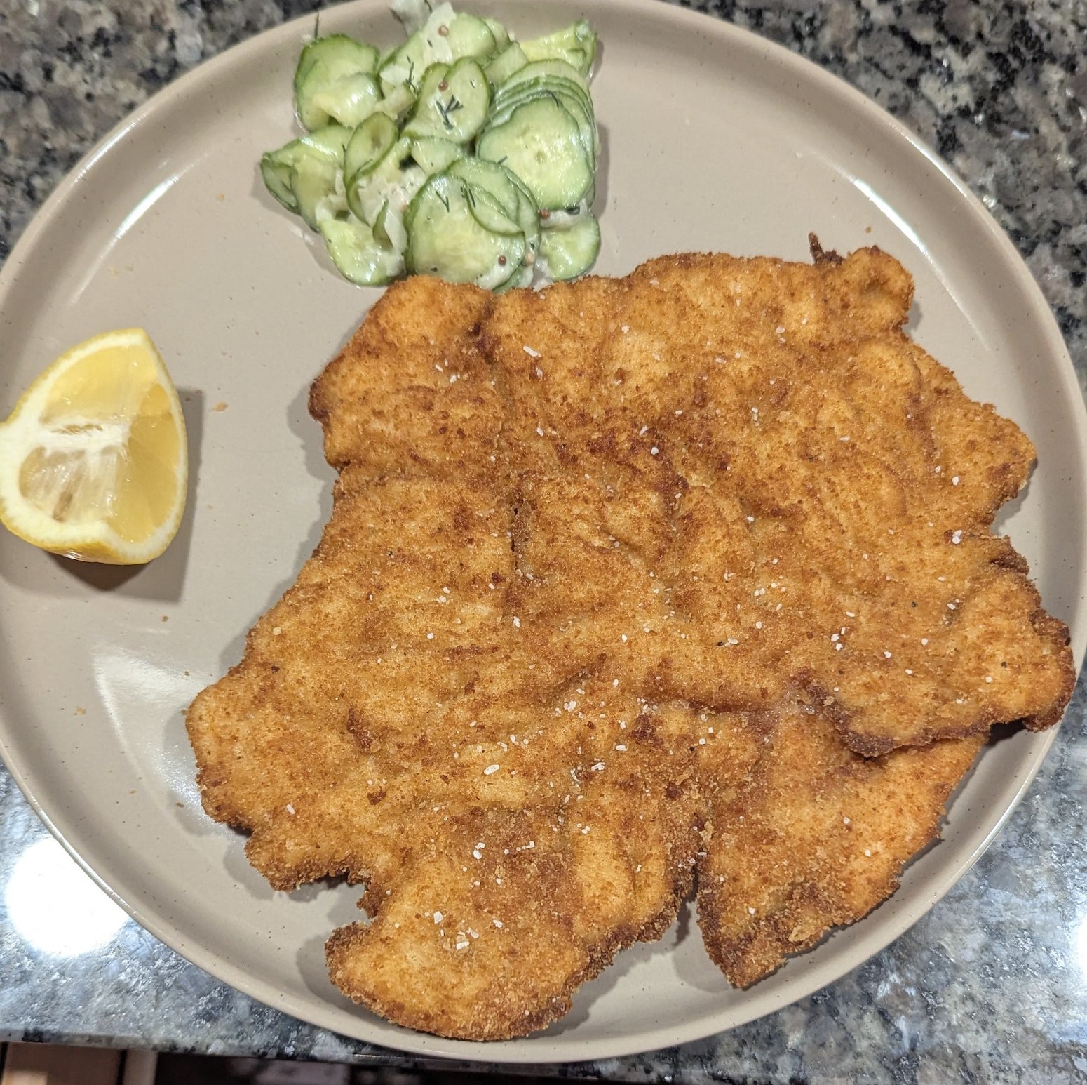 A breaded schnitzel served with a side of cucumber salad and a lemon wedge on a plate.