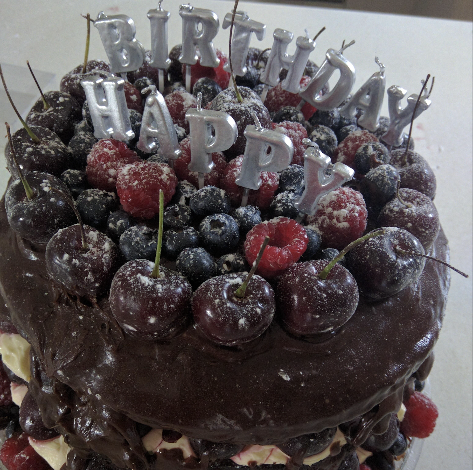 Chocolate cake topped with fresh cherries, raspberries, blueberries, and silver “HAPPY BIRTHDAY” candles.