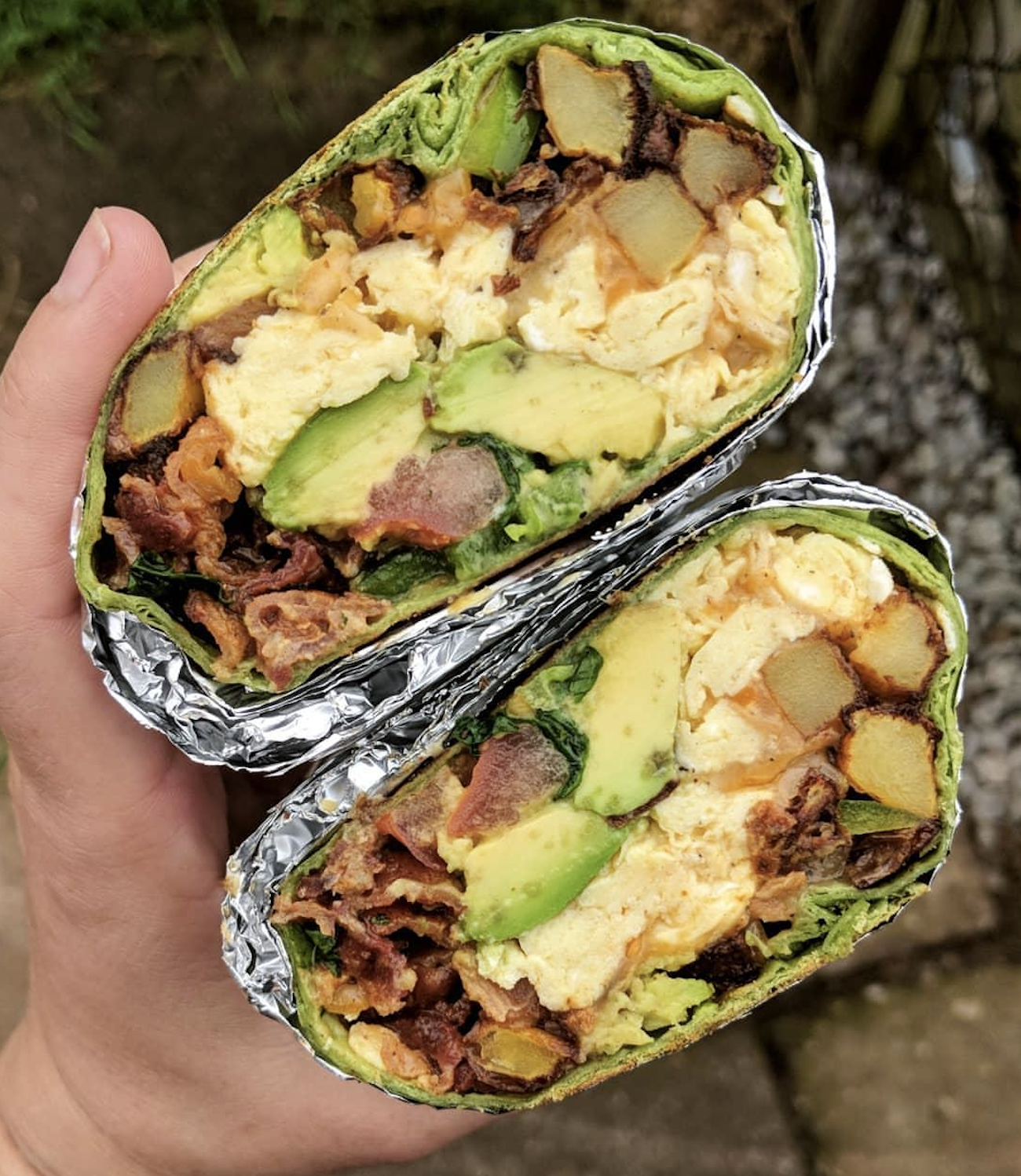 A hand holds two halves of a breakfast burrito wrapped in a green tortilla and foil, filled with scrambled eggs, avocado, potatoes, and bacon.