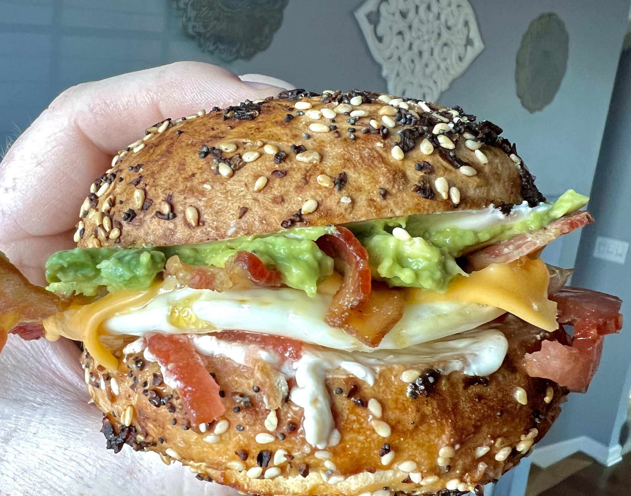 A hand holding an everything bagel breakfast sandwich with bacon, egg, cheese, avocado, and tomato.