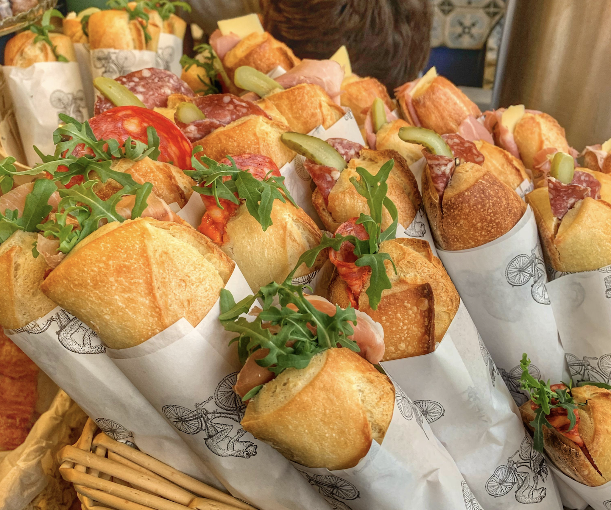 Many baguette sandwiches filled with cured meats, cheese, pickles, and arugula, individually wrapped in printed paper and displayed in a basket.