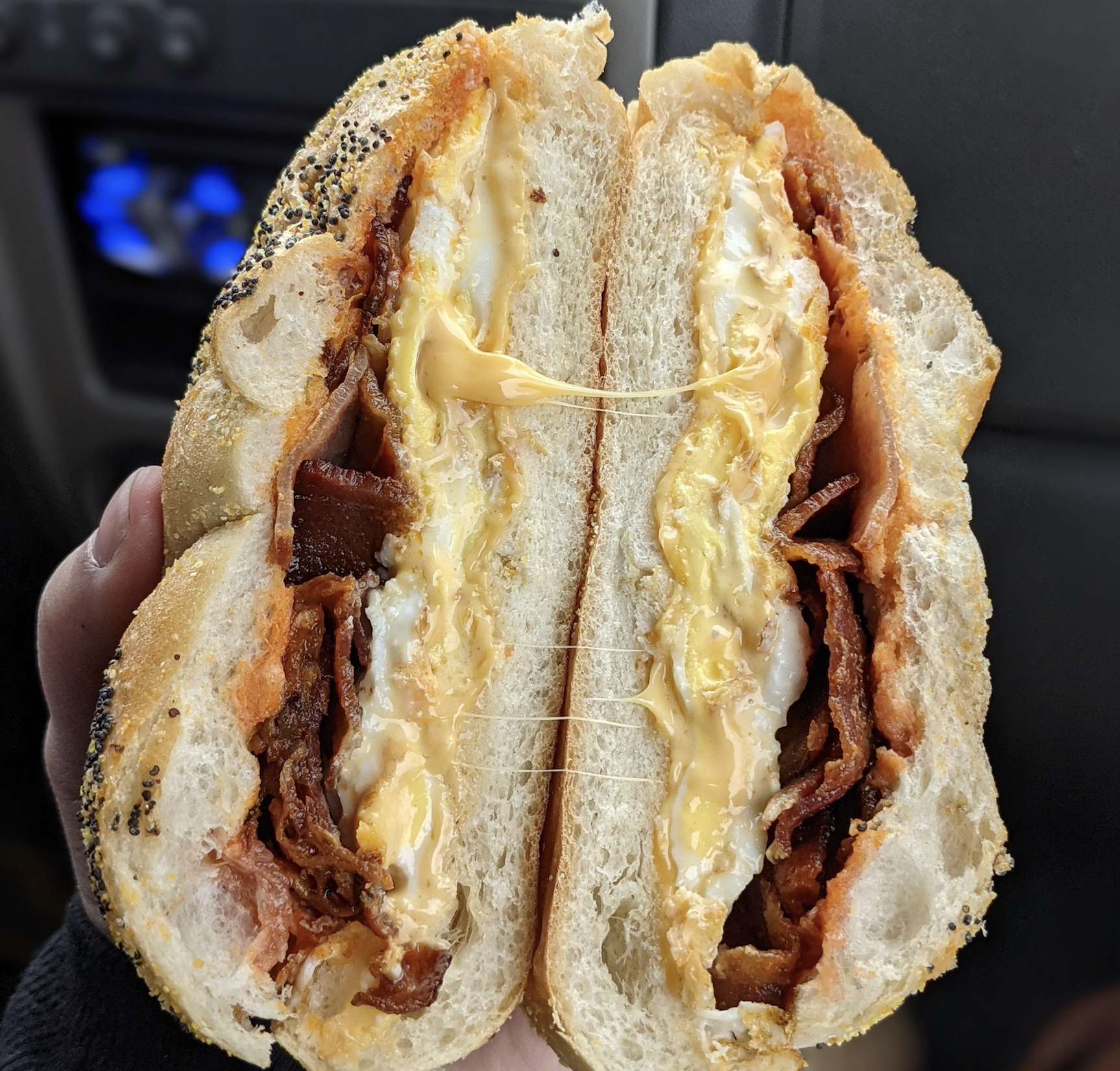 A poppy seed bagel sandwich filled with bacon, egg, and melted cheese, held in two halves with strings of cheese stretching between them.