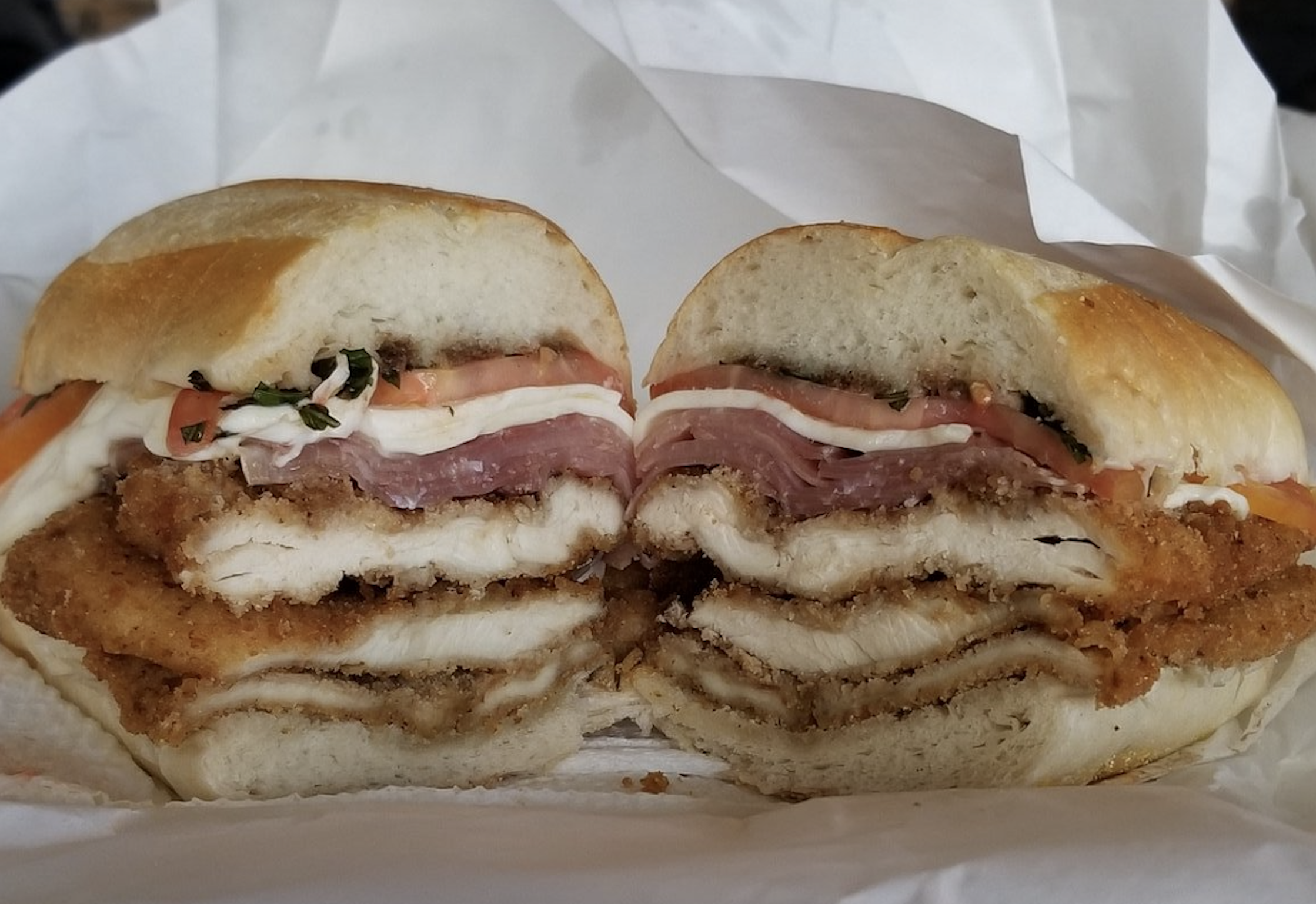 A chicken cutlet sandwich cut in half, layered with ham, fresh mozzarella, and tomato slices on a crusty roll.