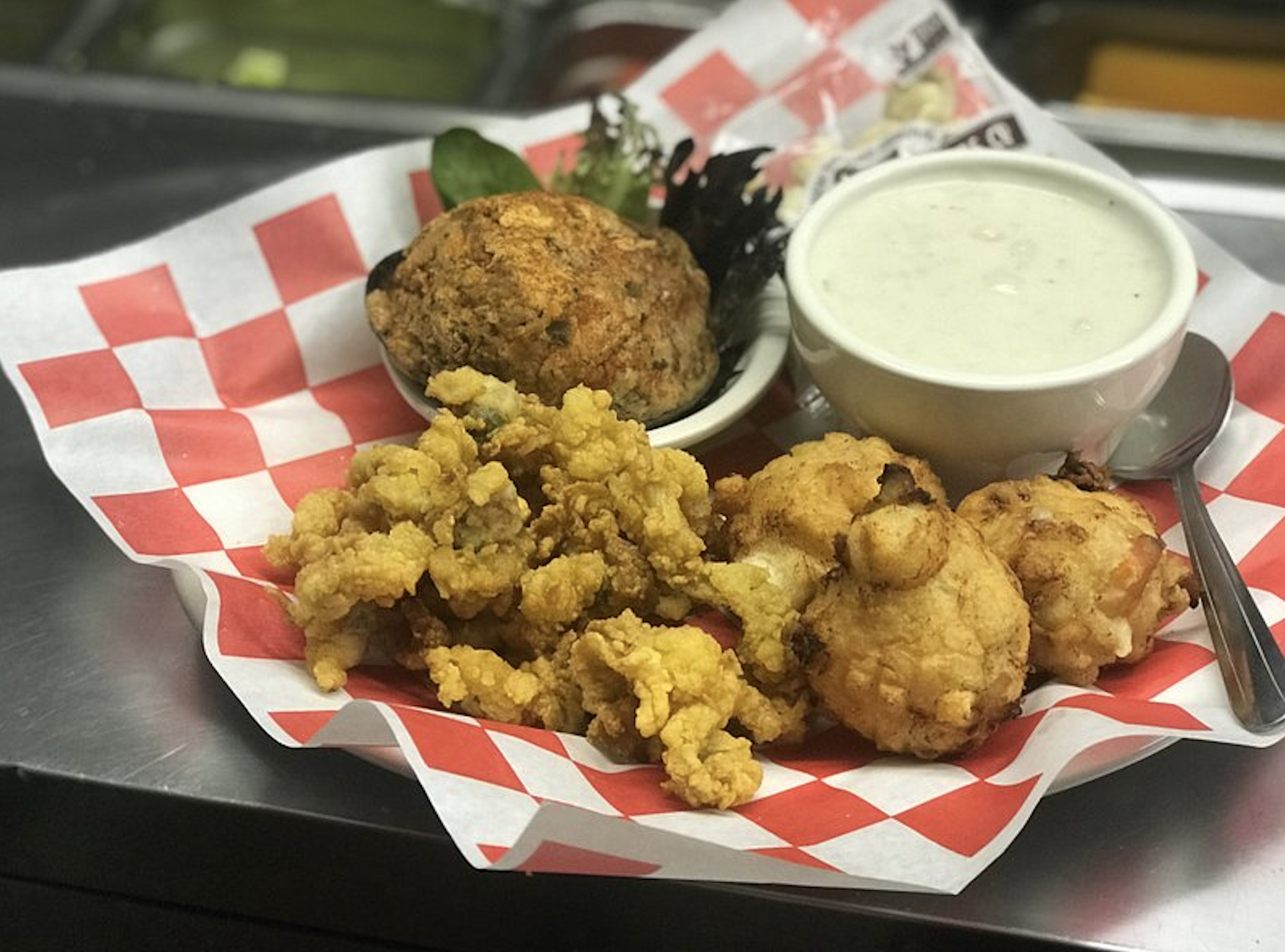 A basket lined with red and white checkered paper containing fried seafood, a large crab cake, and a small bowl of creamy dipping sauce with a spoon.