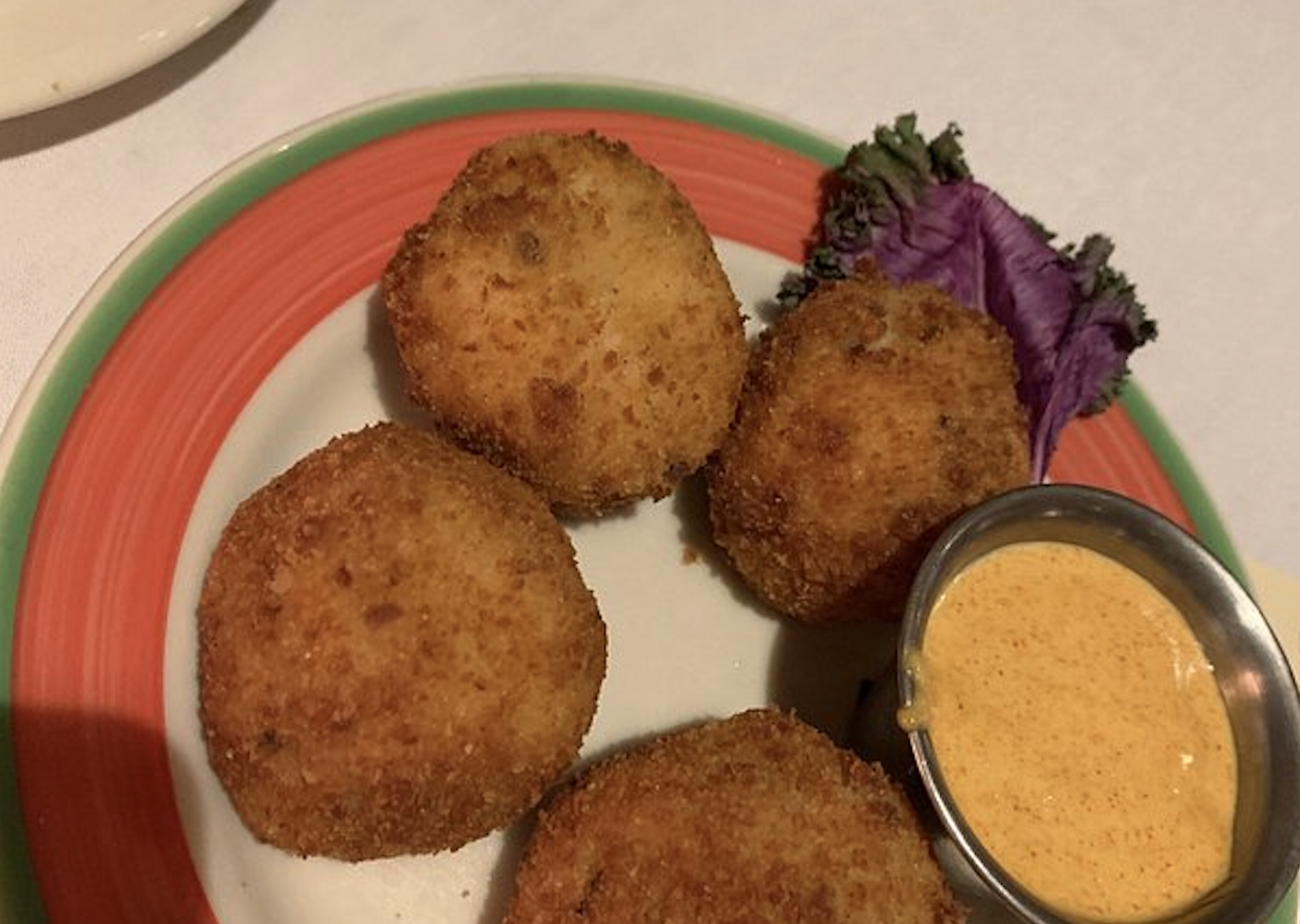 Four breaded, deep-fried appetizer spheres served with a side of dipping sauce and a garnish of purple kale on a decorative plate.