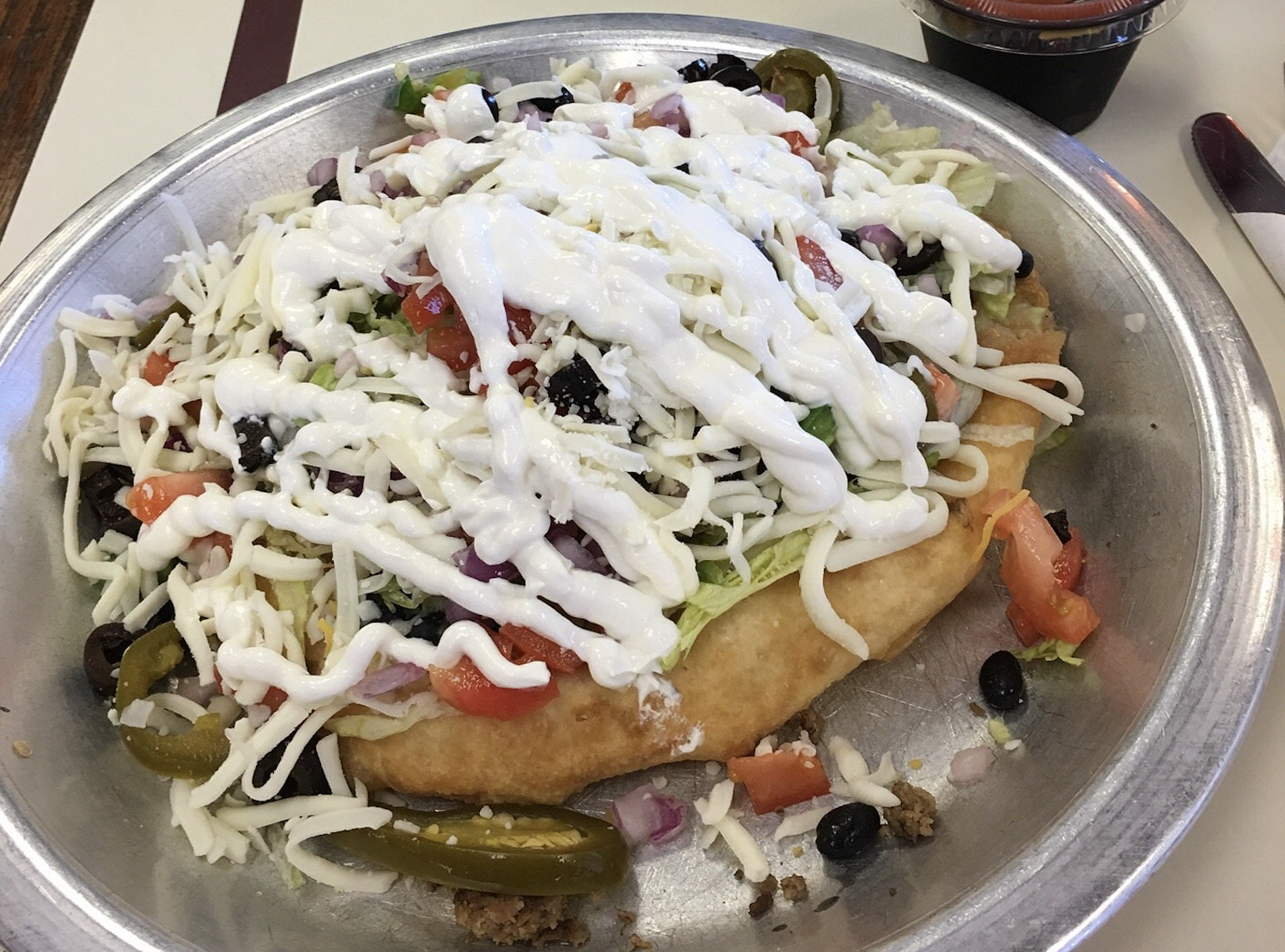Indian taco on fry bread topped with shredded lettuce, diced tomatoes, black olives, jalapeño slices, crumbled cheese, and a drizzle of sour cream.