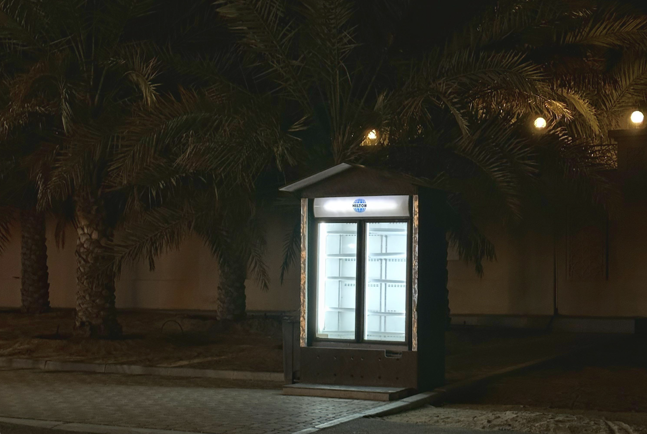 An illuminated, empty outdoor vending fridge with the Hiltons logo stands at night in front of palm trees.
