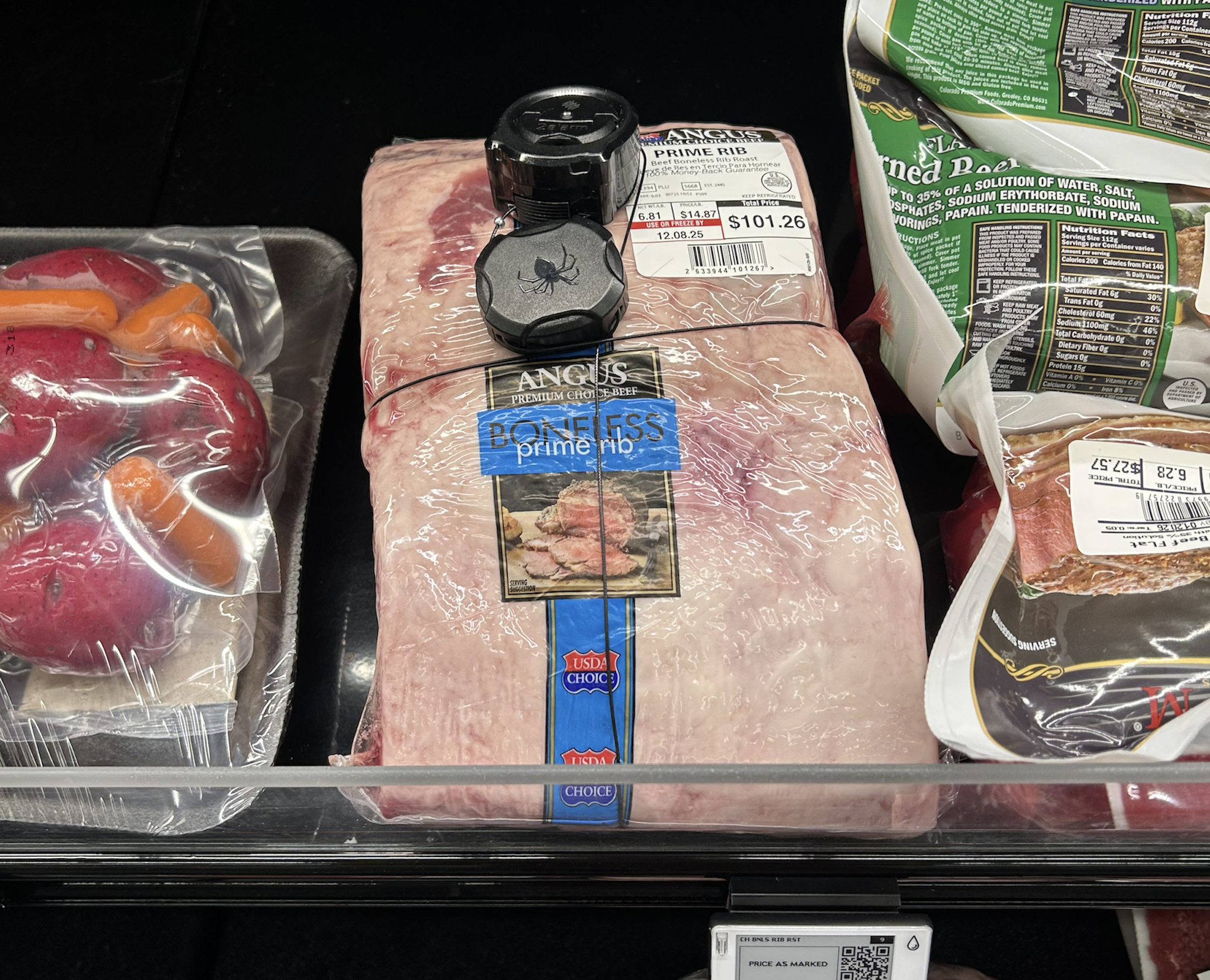 A vacuum-sealed Angus boneless prime rib roast with security tags, priced at $101.26, sitting on a grocery store shelf next to a package of red potatoes.