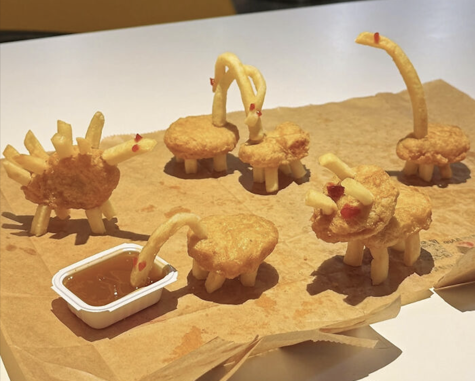 Five dinosaur figures made from chicken nuggets and french fries stand on a piece of parchment paper, with one dinosaur dipping its head into a small container of sauce.