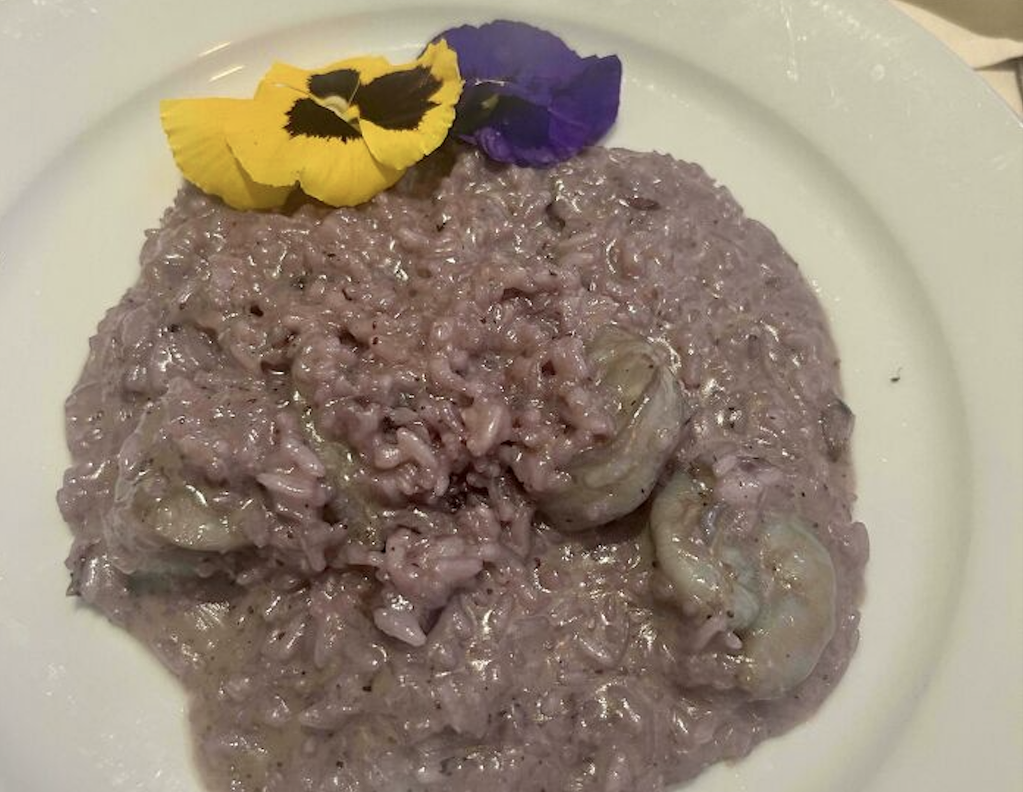Purple risotto with shrimp, garnished with yellow and purple edible flowers on a white plate.