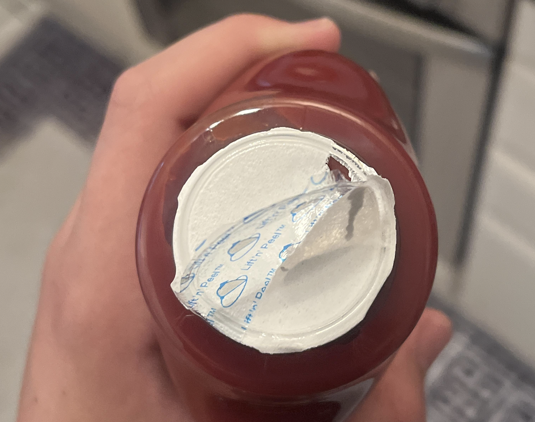A hand holding a bottle of ketchup with a partially peeled foil seal over the opening.