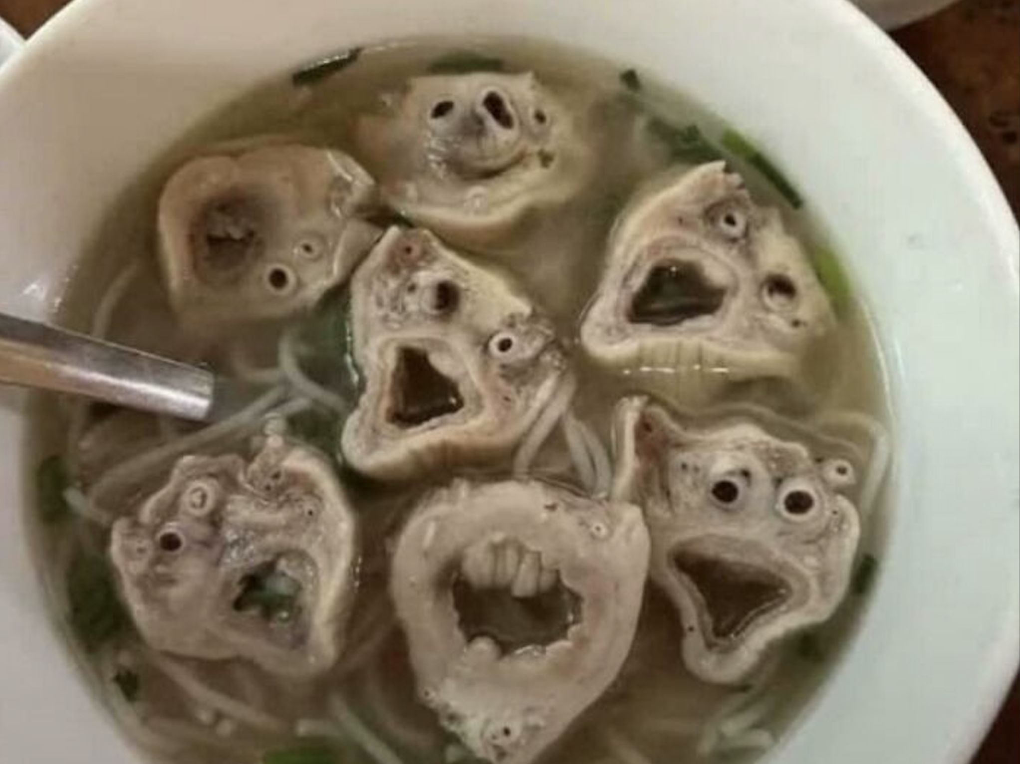 A bowl of noodle soup featuring several pieces of sliced, cooked animal innards with organic shapes that resemble expressive, creature-like faces.