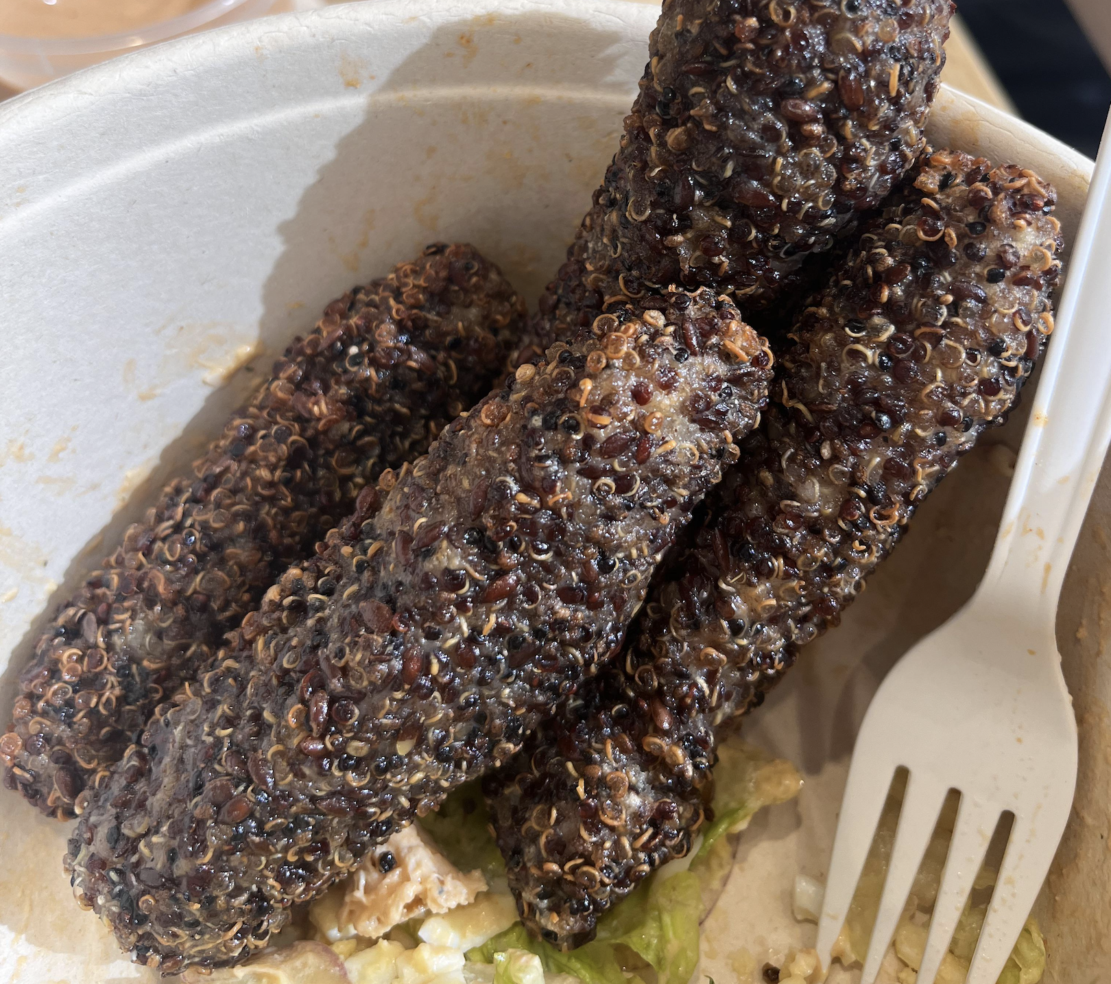 Three sausage-shaped rolls coated in quinoa inside a paper bowl next to a plastic fork.