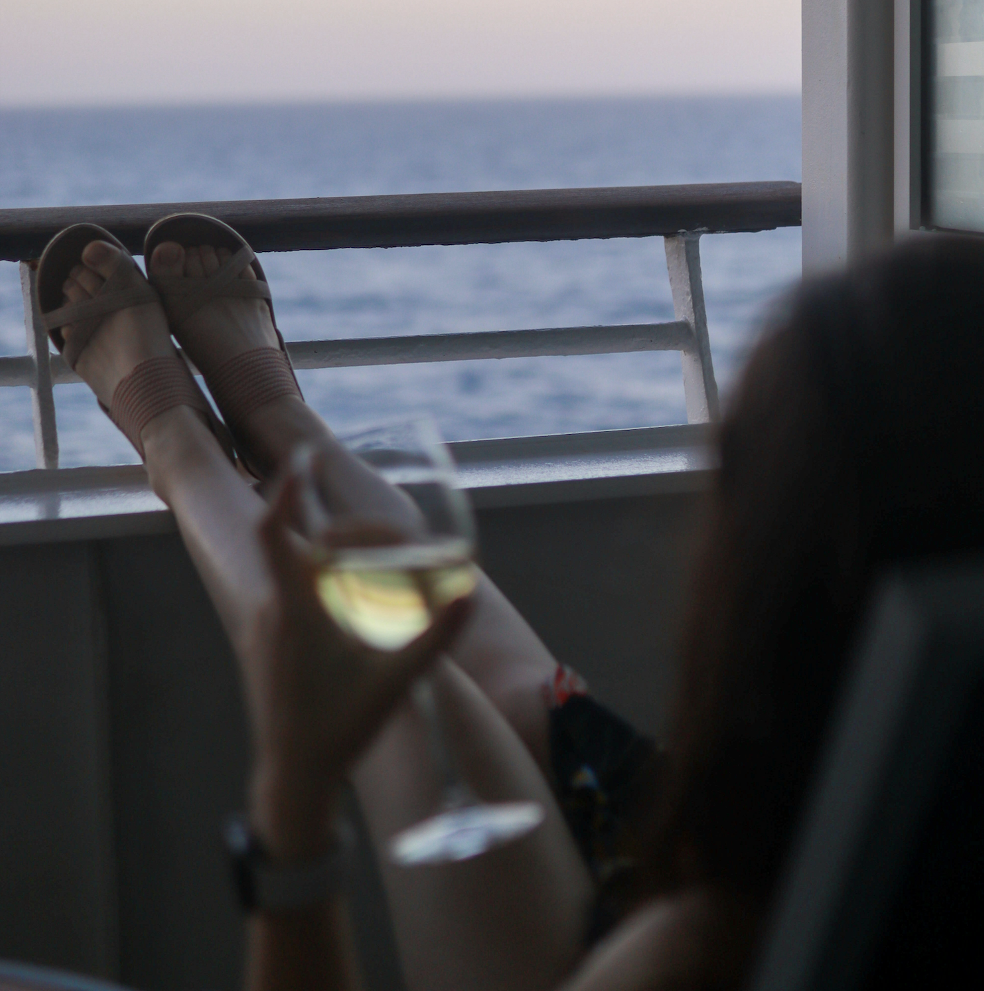 A person relaxing on a balcony with their feet up on the railing, holding a glass of white wine while enjoying the cruise entertainment and ocean view in the background.