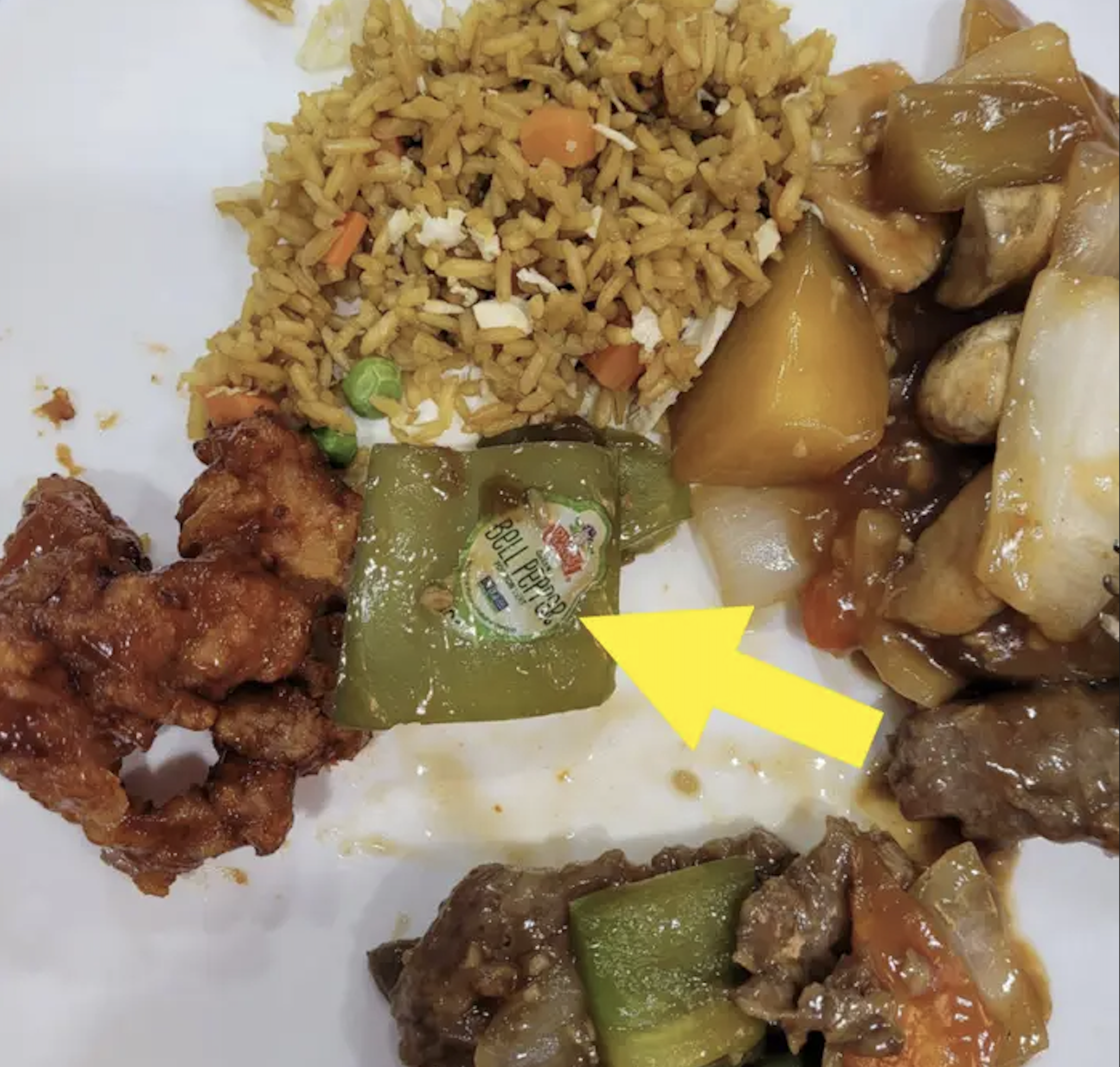 A plate of Chinese food featuring fried rice, chicken, and stir-fried vegetables, with a yellow arrow pointing to a piece of green bell pepper that still has its produce sticker attached.