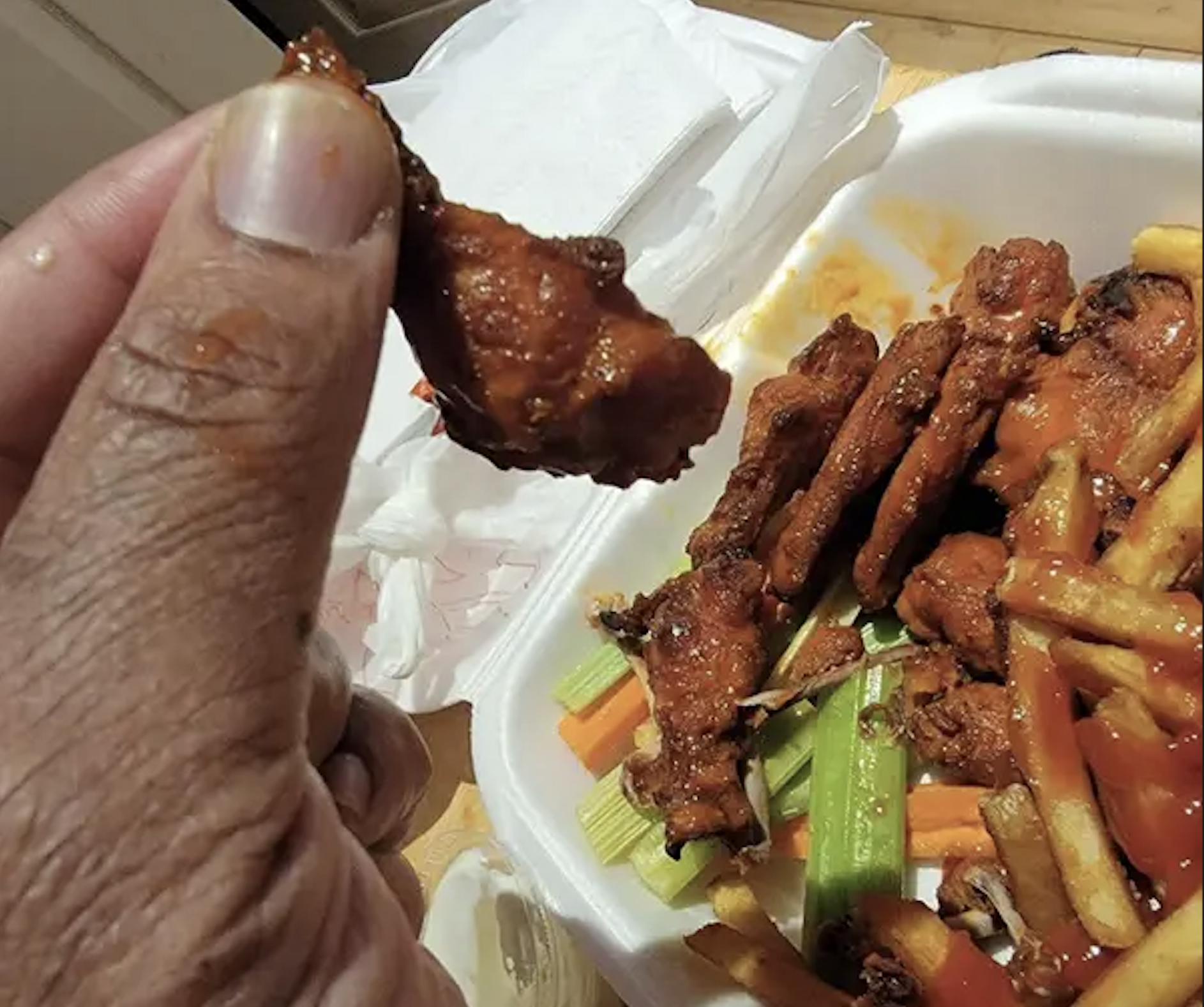 A hand holding a single buffalo wing above a takeout container filled with more wings, french fries, celery, and carrots.