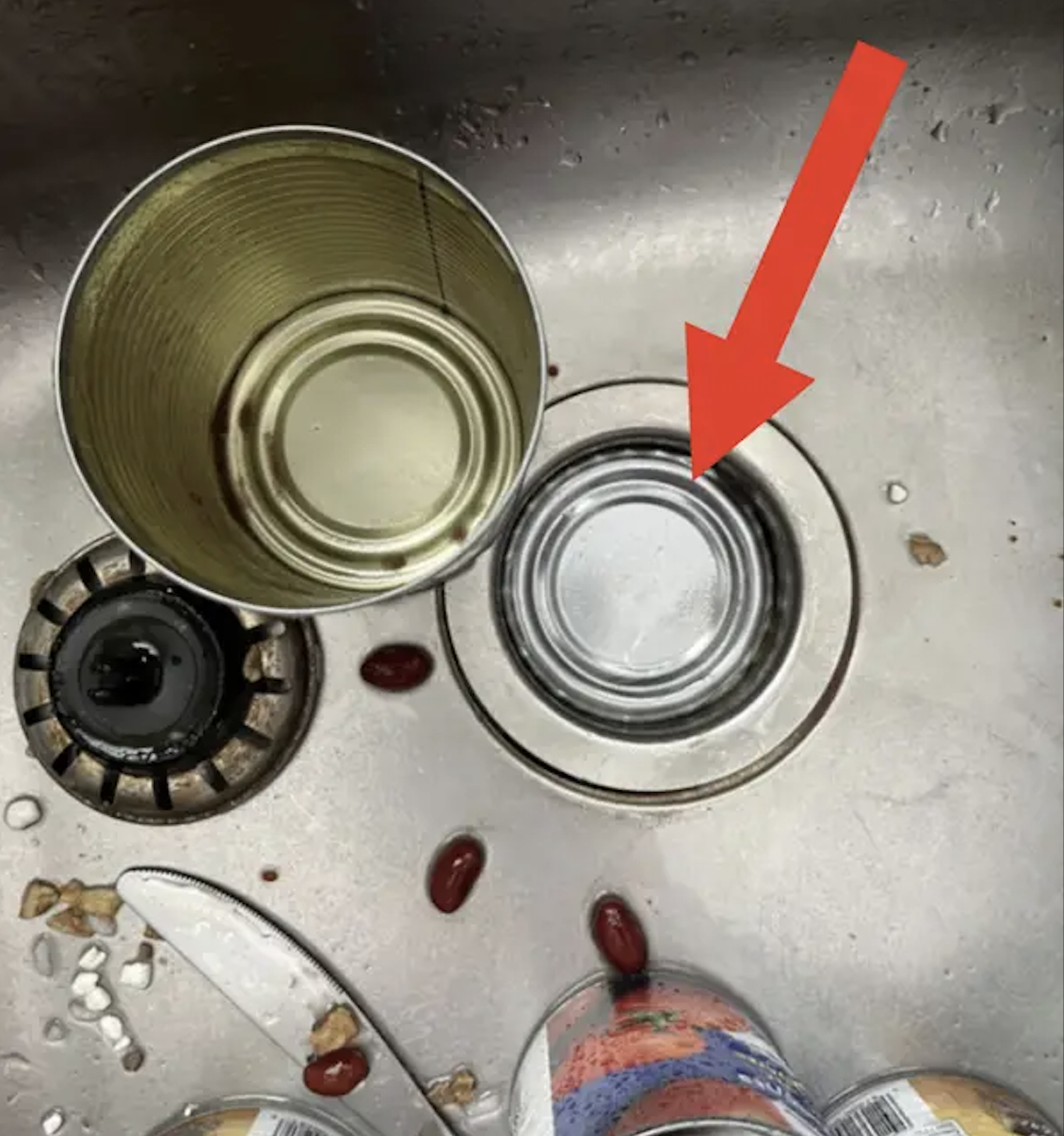 A red arrow points to a metal can lid resting inside a kitchen sink drain.