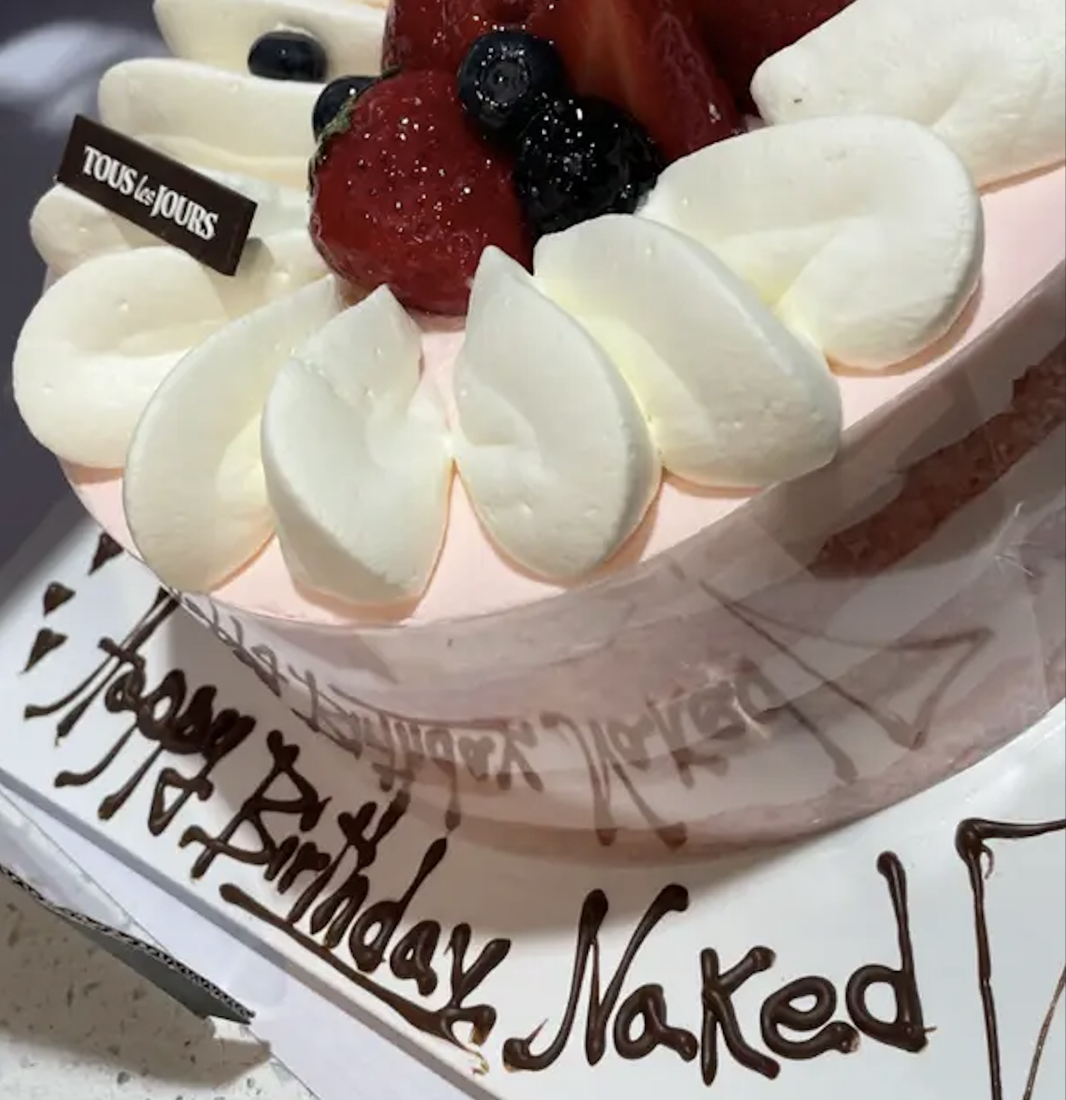 A round pink cake from Tous les Jours topped with whipped cream and berries, with “Happy Birthday Naked” written in chocolate on the base.