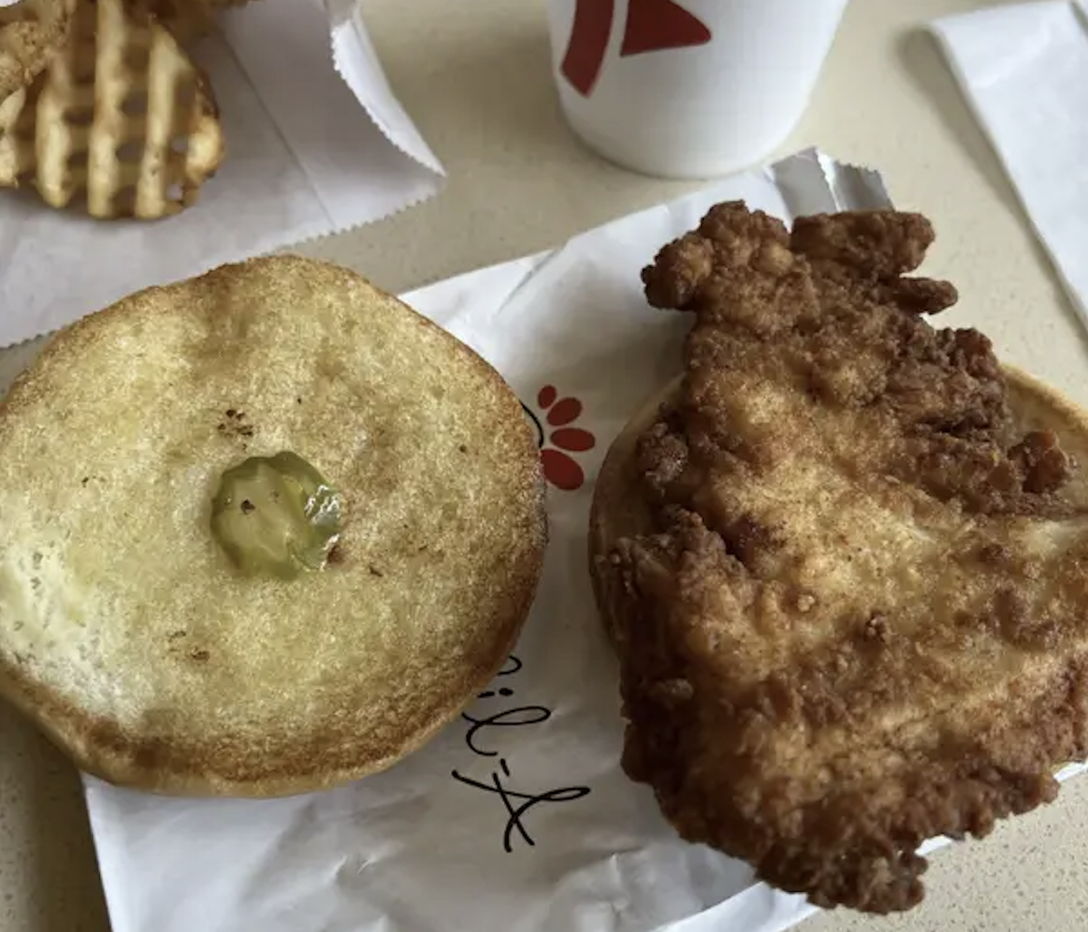 An open Chick-fil-A chicken sandwich with a single pickle on the bun, next to waffle fries and a drink.