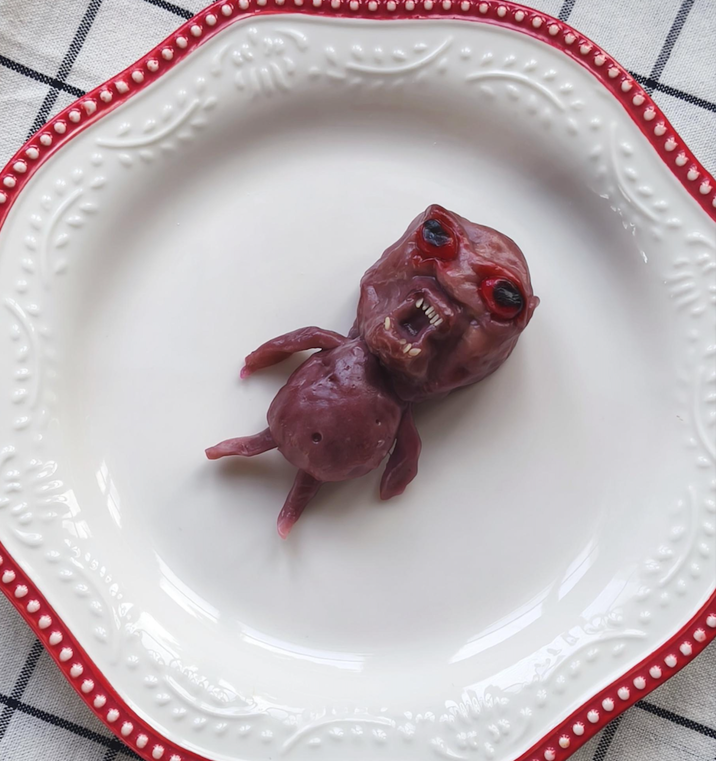 A small, dark red, humanoid creature figurine with a snarling face and black eyes resting on a white plate with a decorative red rim.