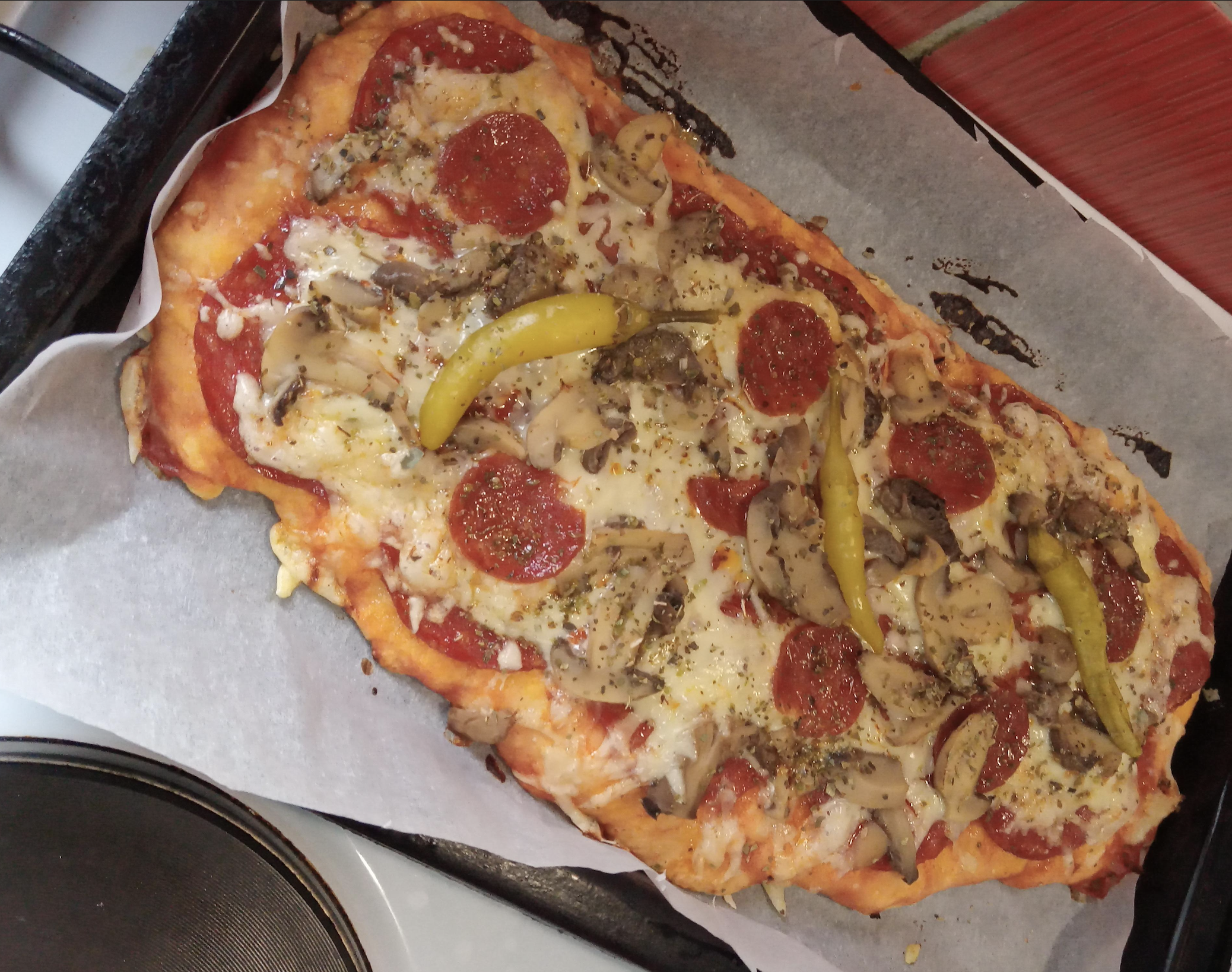 Rectangular flatbread pizza topped with pepperoni, mushrooms, and three yellow chili peppers on a baking sheet.