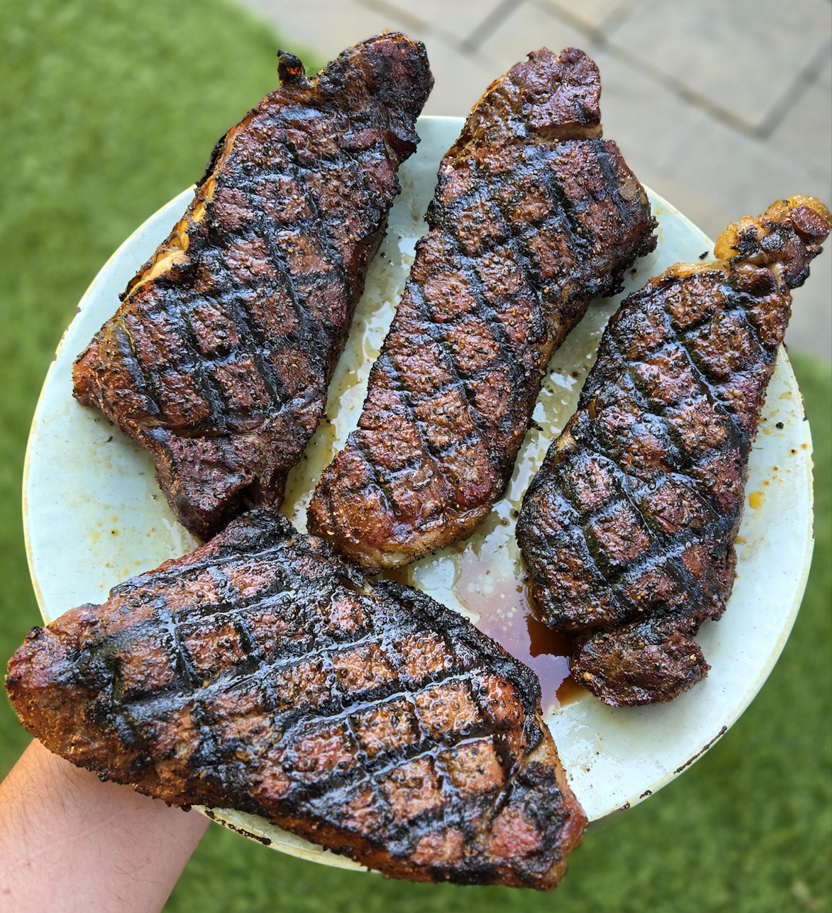 Four grilled steaks with distinct sear marks on a plate held by a hand.