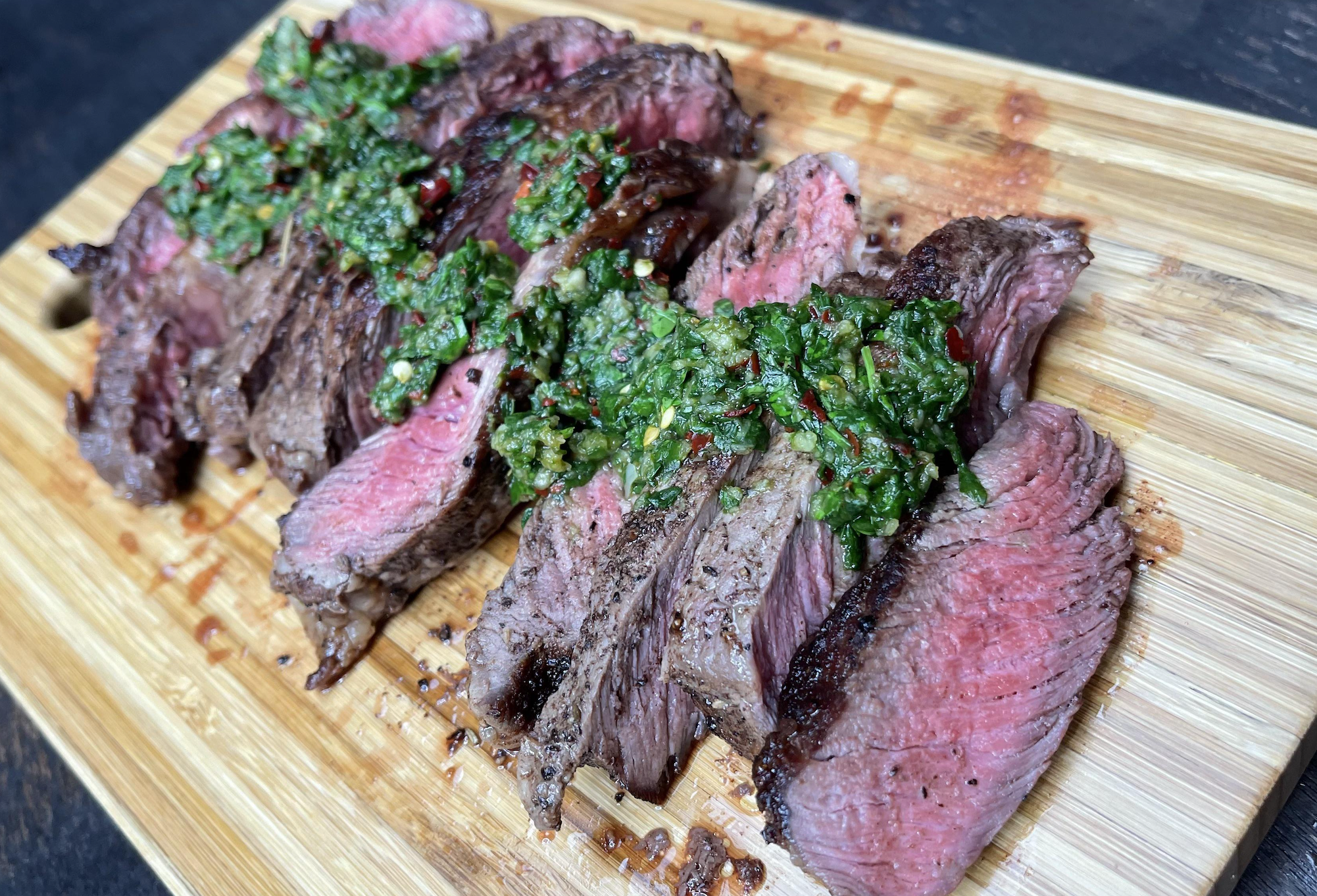 Sliced medium-rare steak topped with chimichurri sauce on a wooden cutting board.