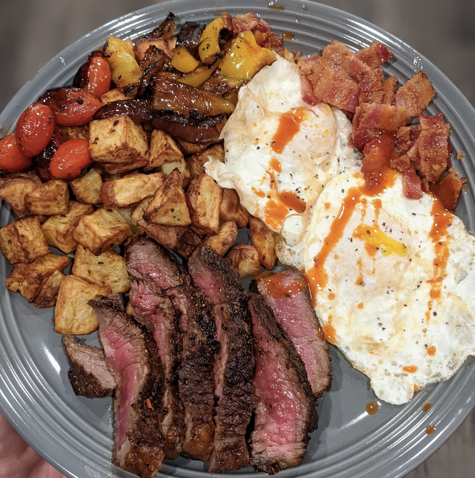 A plate of breakfast featuring sliced steak, two fried eggs drizzled with hot sauce, crispy bacon, roasted potatoes, and blistered tomatoes with peppers.