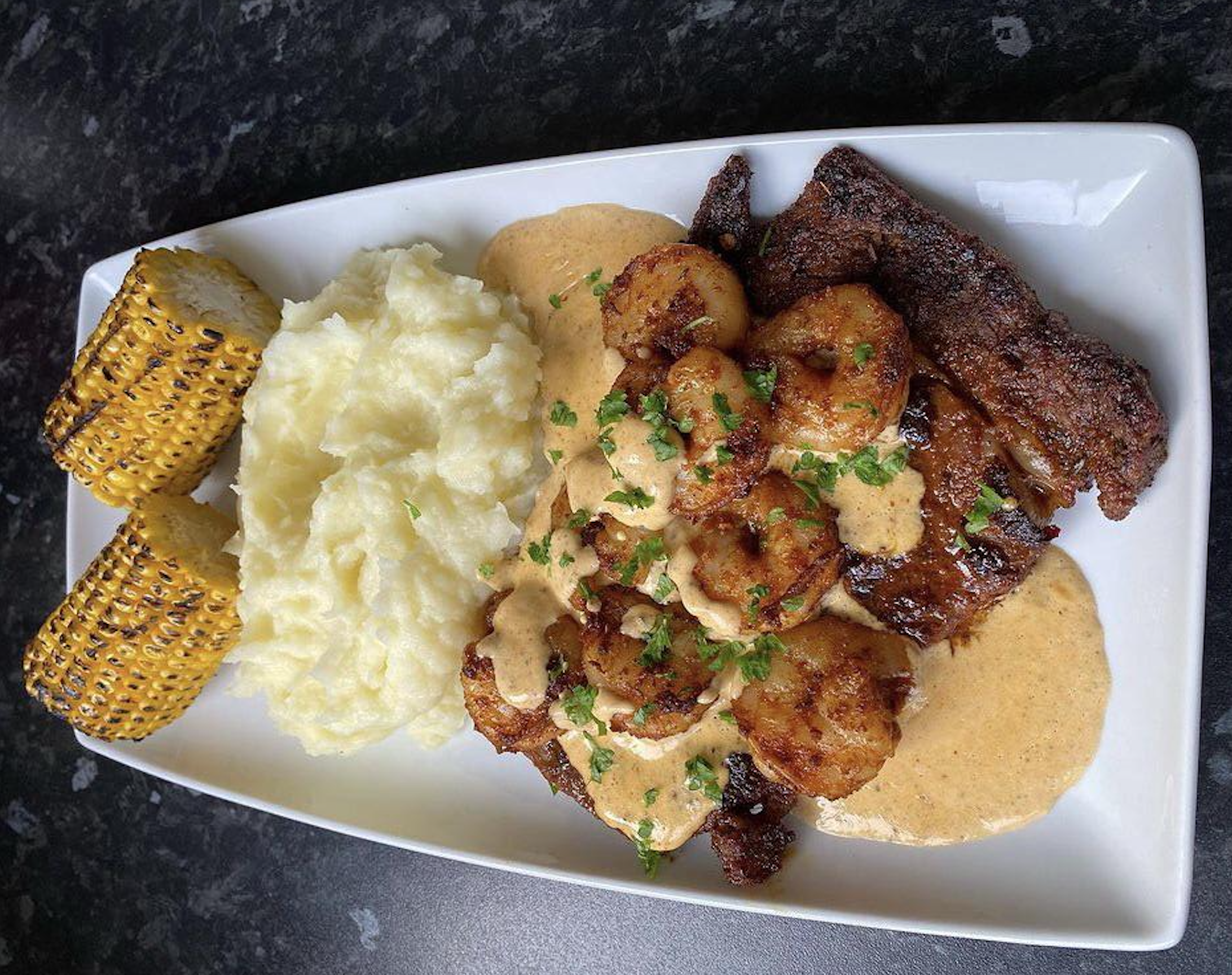 A white rectangular plate featuring a serving of steak topped with shrimp in a creamy sauce, accompanied by mashed potatoes and two pieces of corn on the cob.
