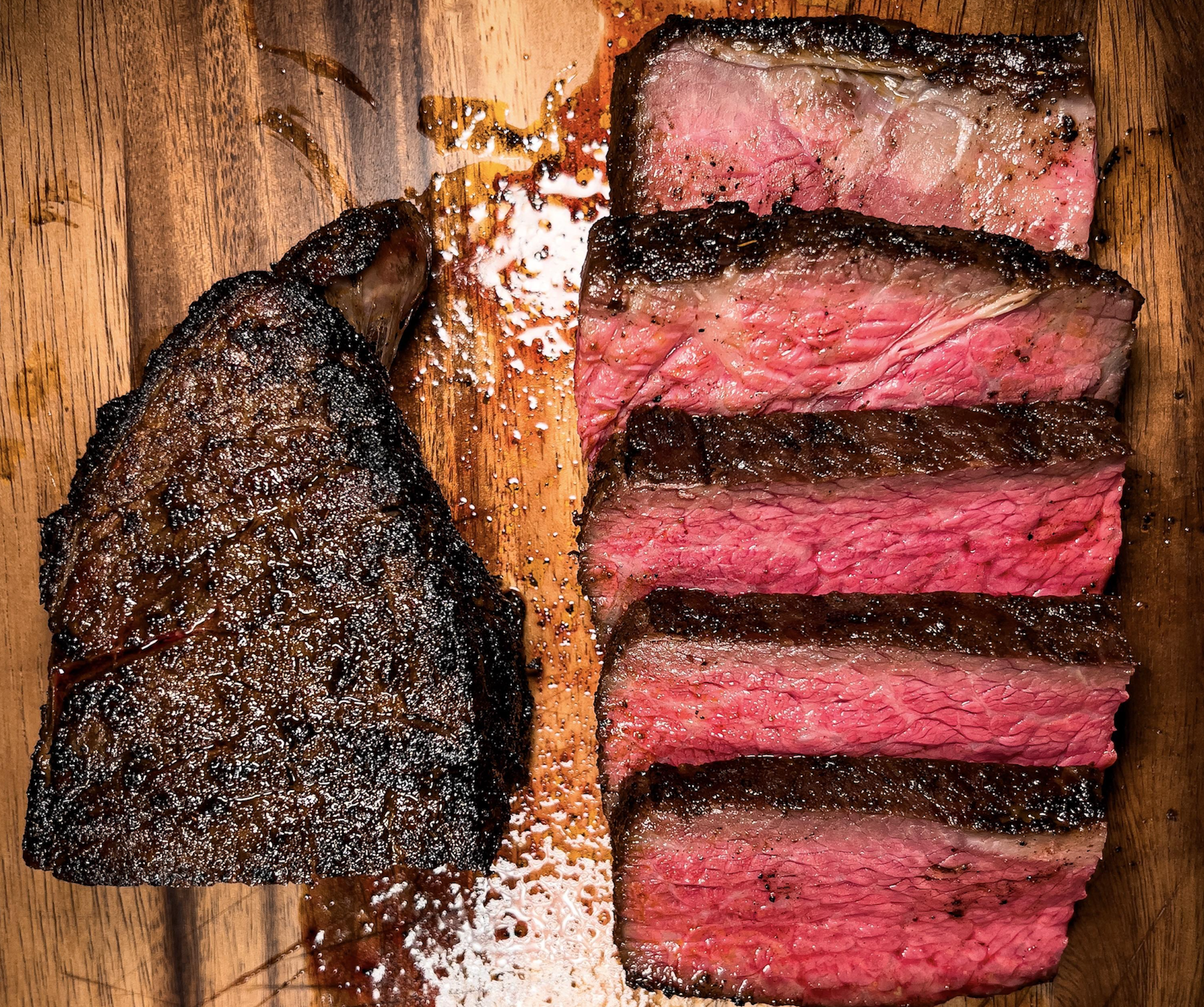 A large seared steak sits next to five thick, medium-rare slices on a wooden cutting board.
