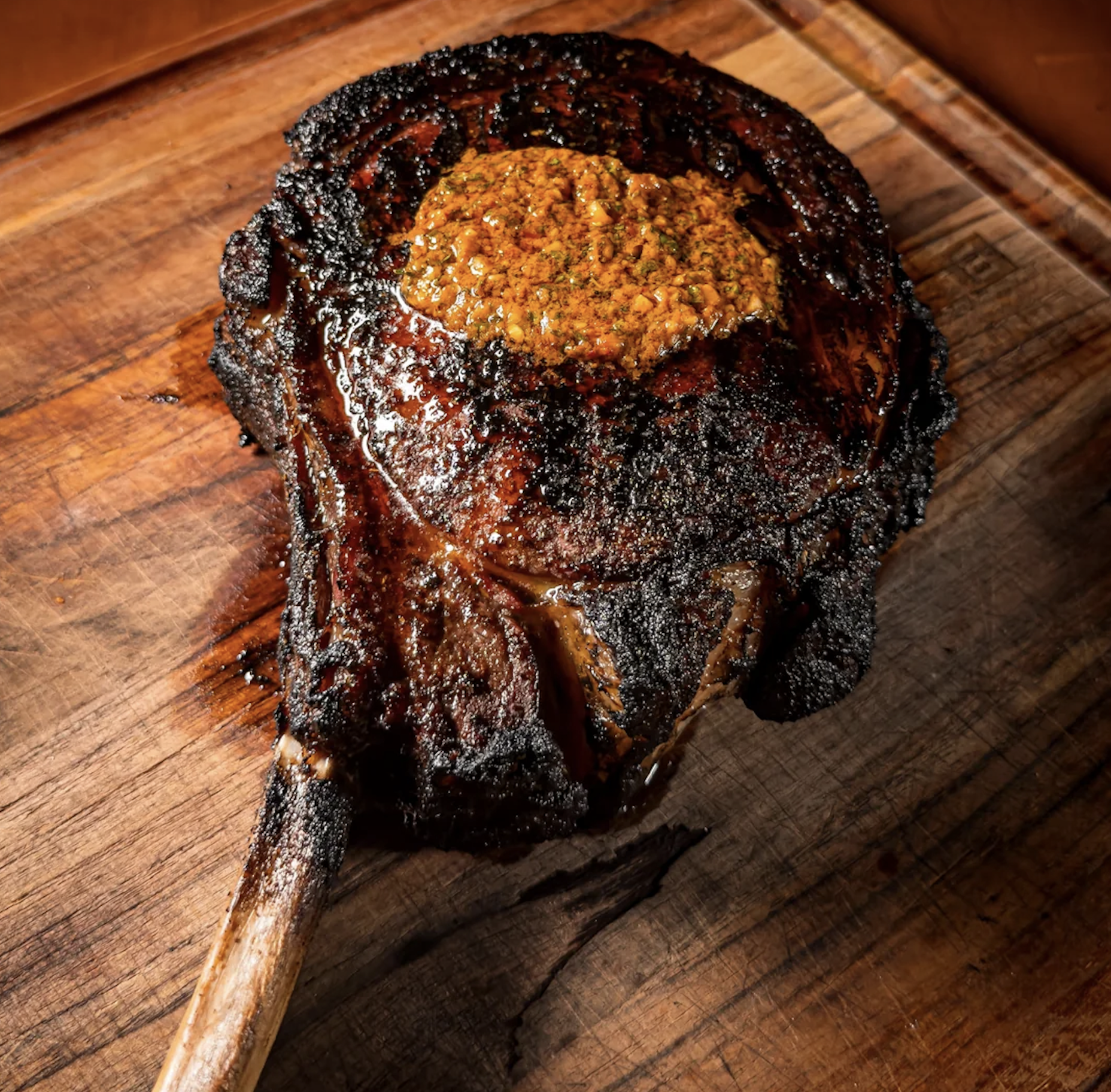 A charred tomahawk steak topped with a dollop of herb butter on a wooden cutting board.