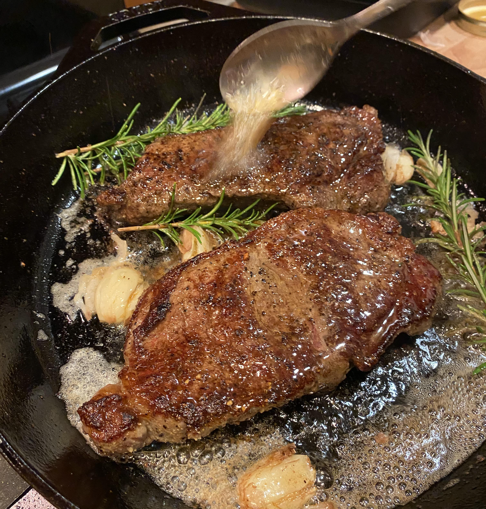Two steaks searing in a cast-iron skillet with rosemary sprigs and garlic cloves, being basted with butter from a spoon.