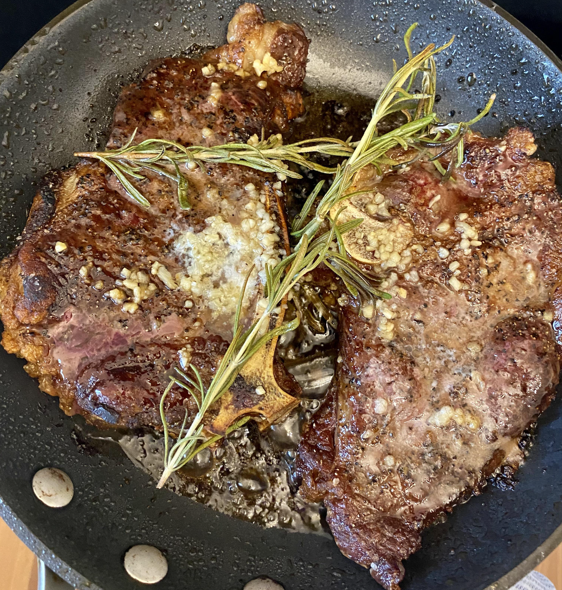 Two steaks searing in a skillet with butter, minced garlic, and fresh rosemary sprigs.
