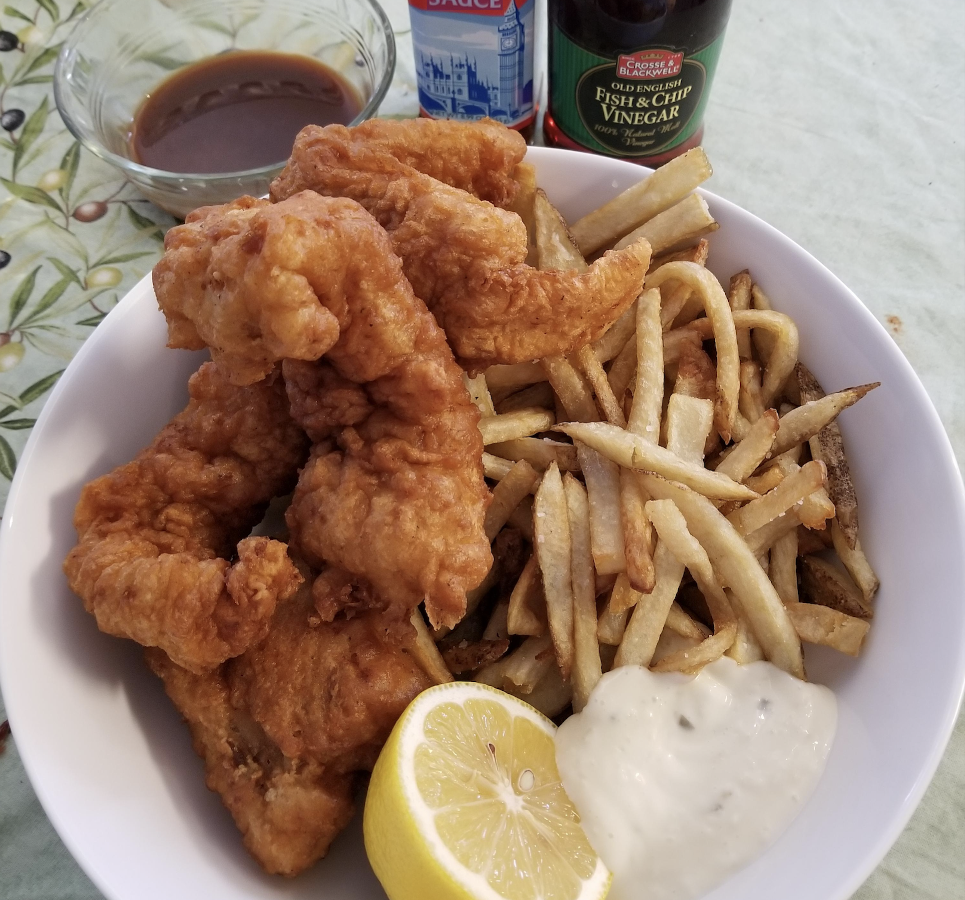 A bowl of fried fish and french fries with a lemon wedge, tartar sauce, a small dish of gravy, and bottles of vinegar and sauce.