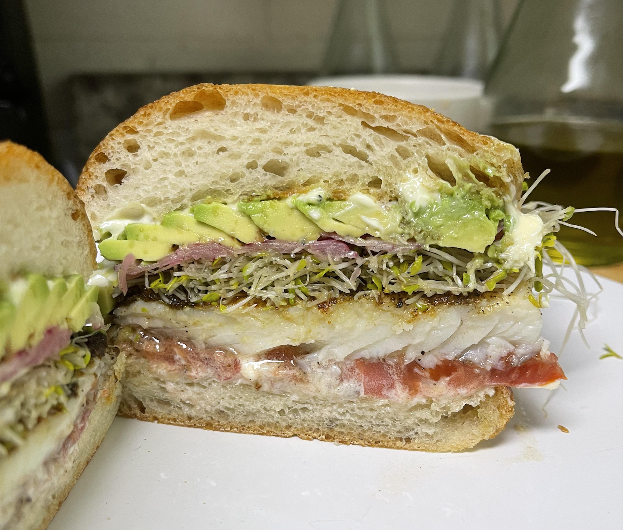 A cross-section of a sandwich on crusty bread layered with avocado, sprouts, red onion, white fish, and tomato.