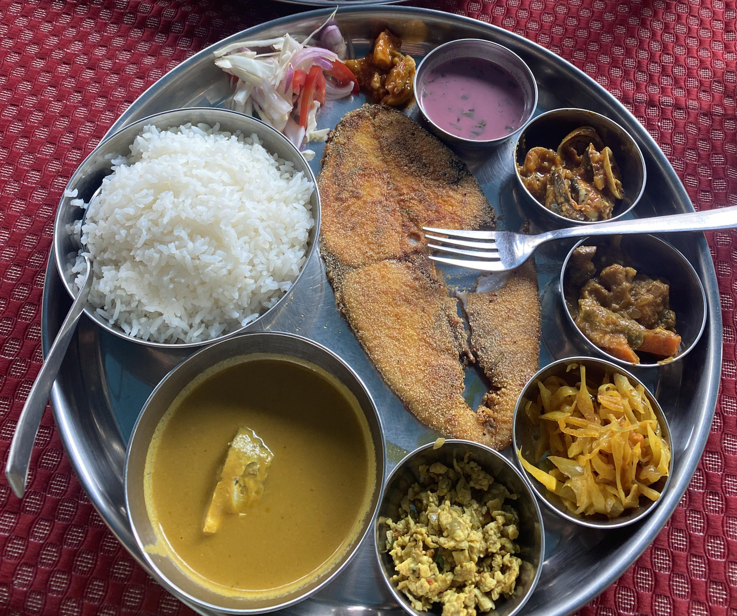 A metal platter holding a large piece of fried fish, a mound of white rice, a bowl of yellow curry, and several small bowls containing vegetable sides, pickles, and a yogurt drink.