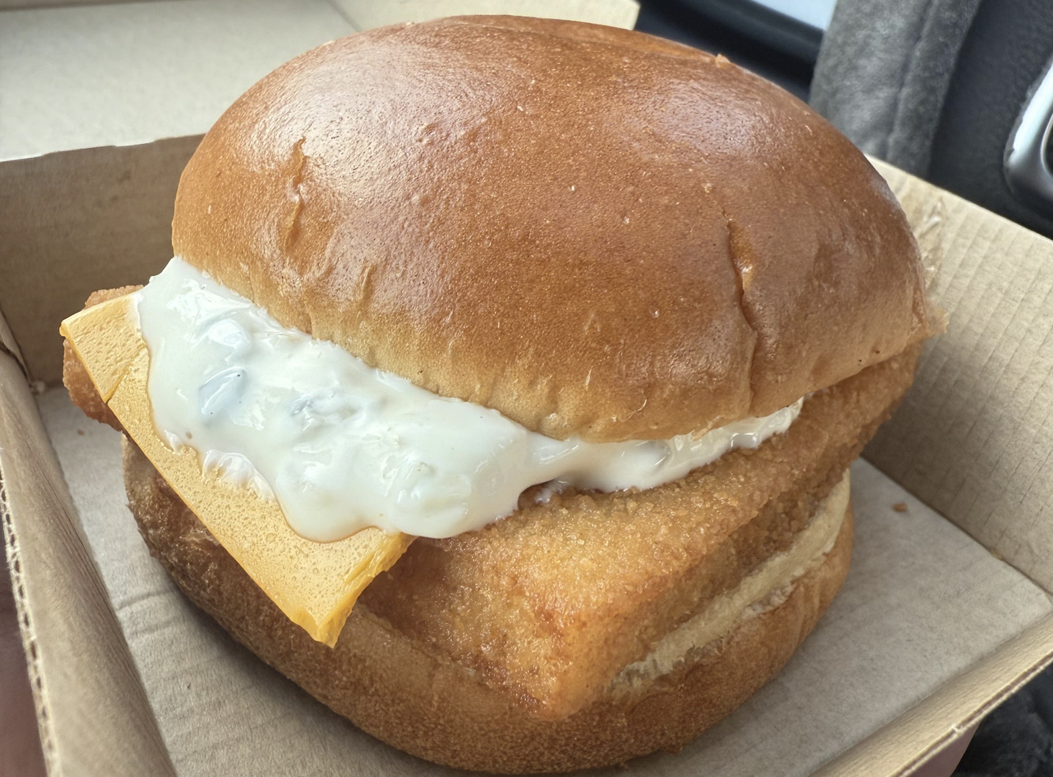 A fish sandwich on a toasted bun with a slice of yellow cheese and a generous dollop of tartar sauce, served in a cardboard box.