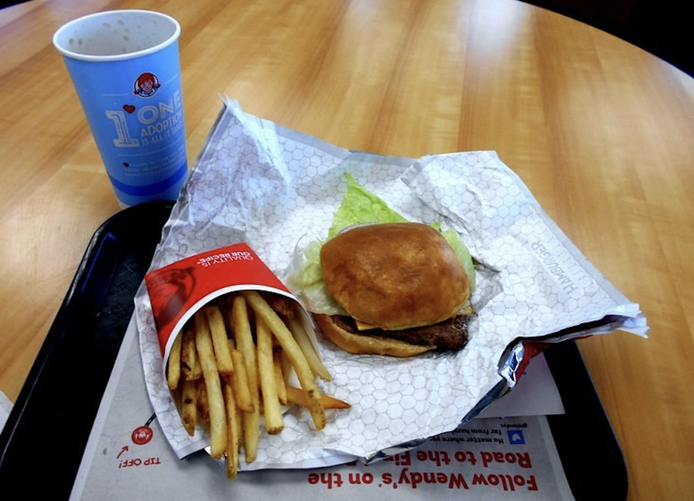 A Wendy’s meal consisting of a cheeseburger, a carton of french fries, and a paper cup on a tray.