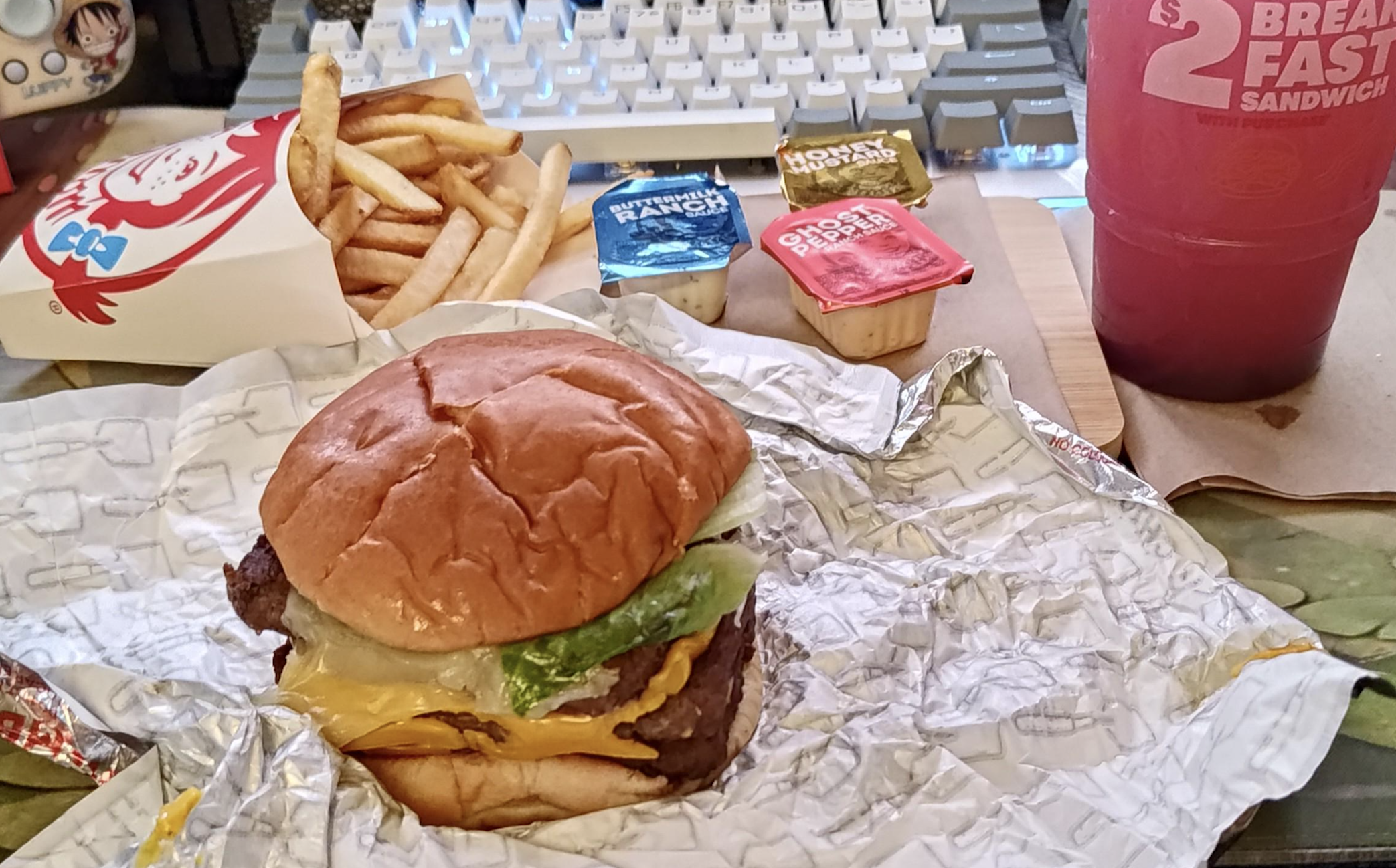 A cheeseburger in a foil wrapper, a container of fries, three dipping sauce cups, and a drink, sitting on a desk in front of a computer keyboard.
