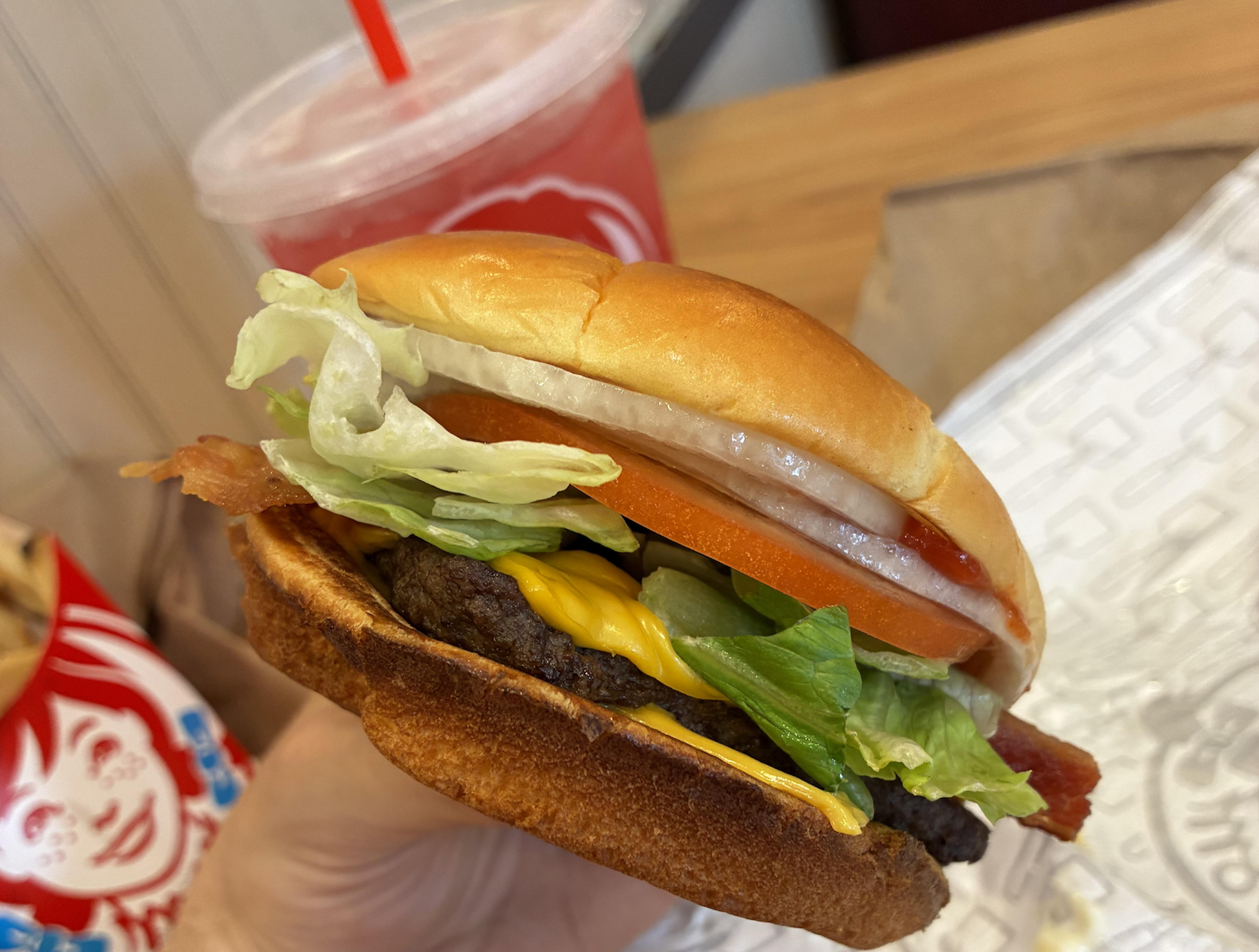 A hand holding a bacon cheeseburger with lettuce, tomato, and onion, with a red drink in the background.