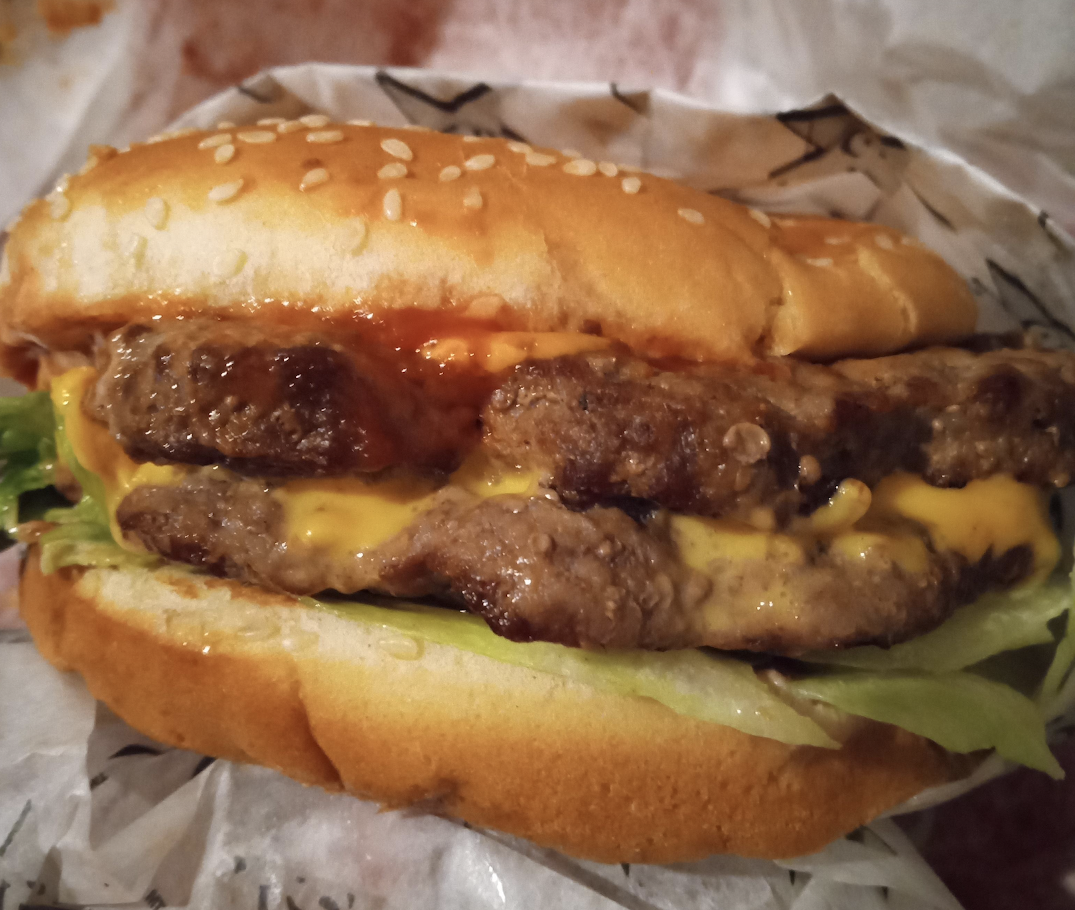 A double cheeseburger with lettuce and ketchup on a sesame seed bun, wrapped in patterned paper.